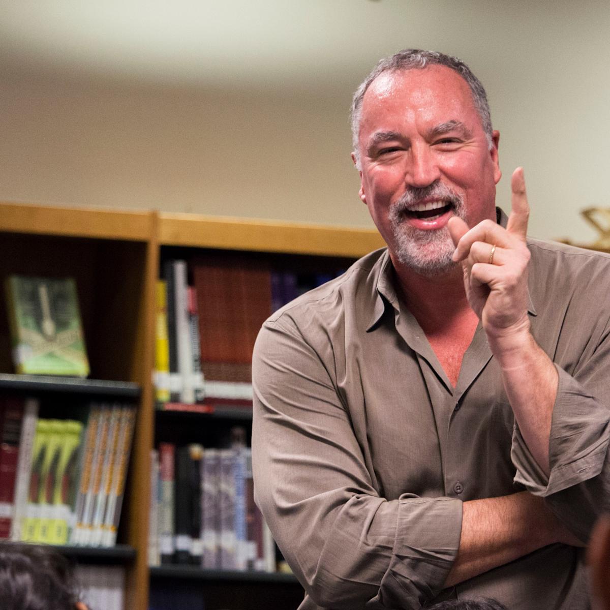 A middle-aged man with gray hair and beard wearing a tan button-down shirt smiles and gestures animatedly while sitting in an office with bookshelves in the background. The candid portrait captures him mid-conversation in a relaxed professional setting.