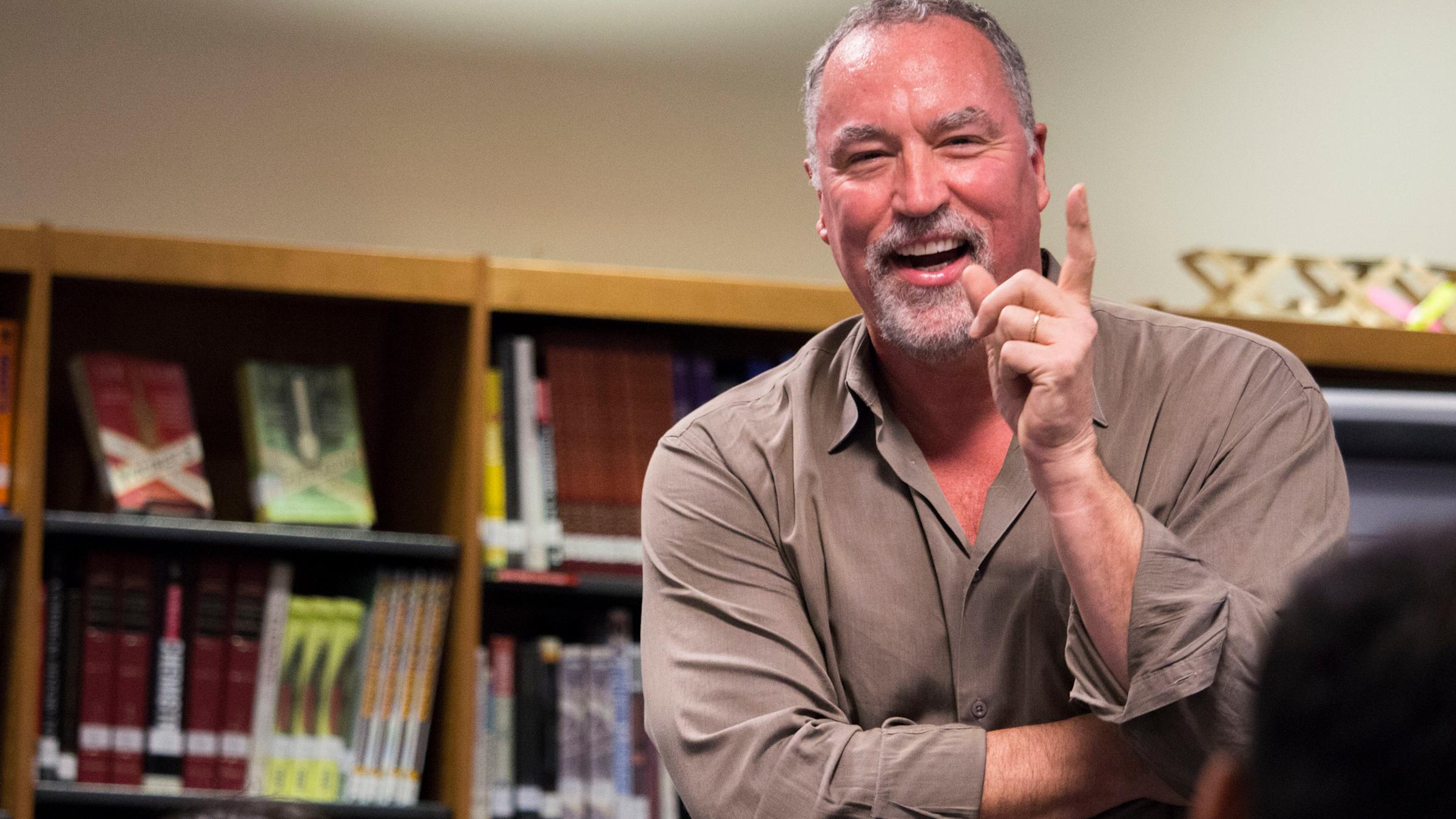 A middle-aged man with gray hair and beard wearing a tan button-down shirt smiles and gestures animatedly while sitting in an office with bookshelves in the background. The candid portrait captures him mid-conversation in a relaxed professional setting.