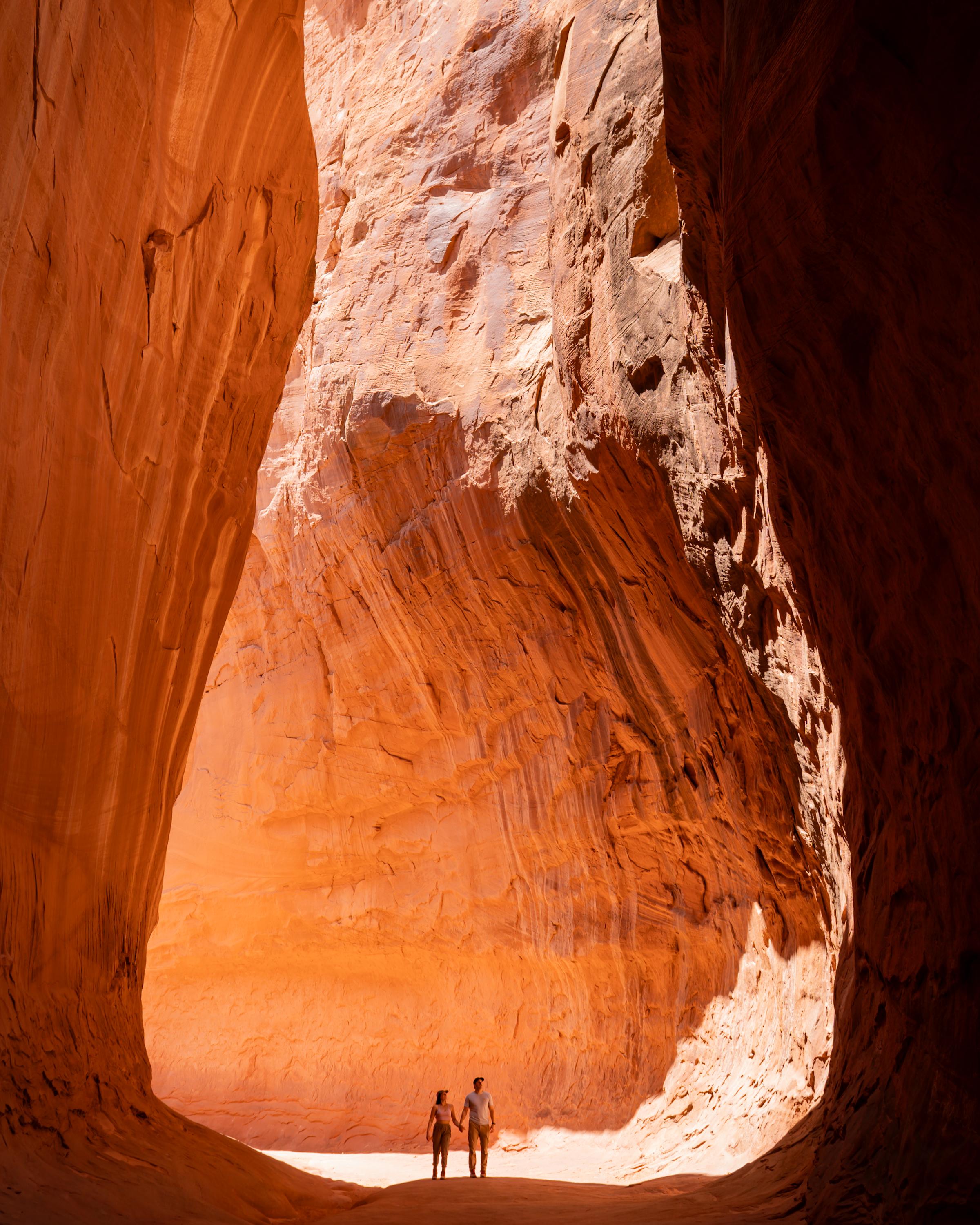 Man and a woman walk in a canyon, beneath a huge rock formations.