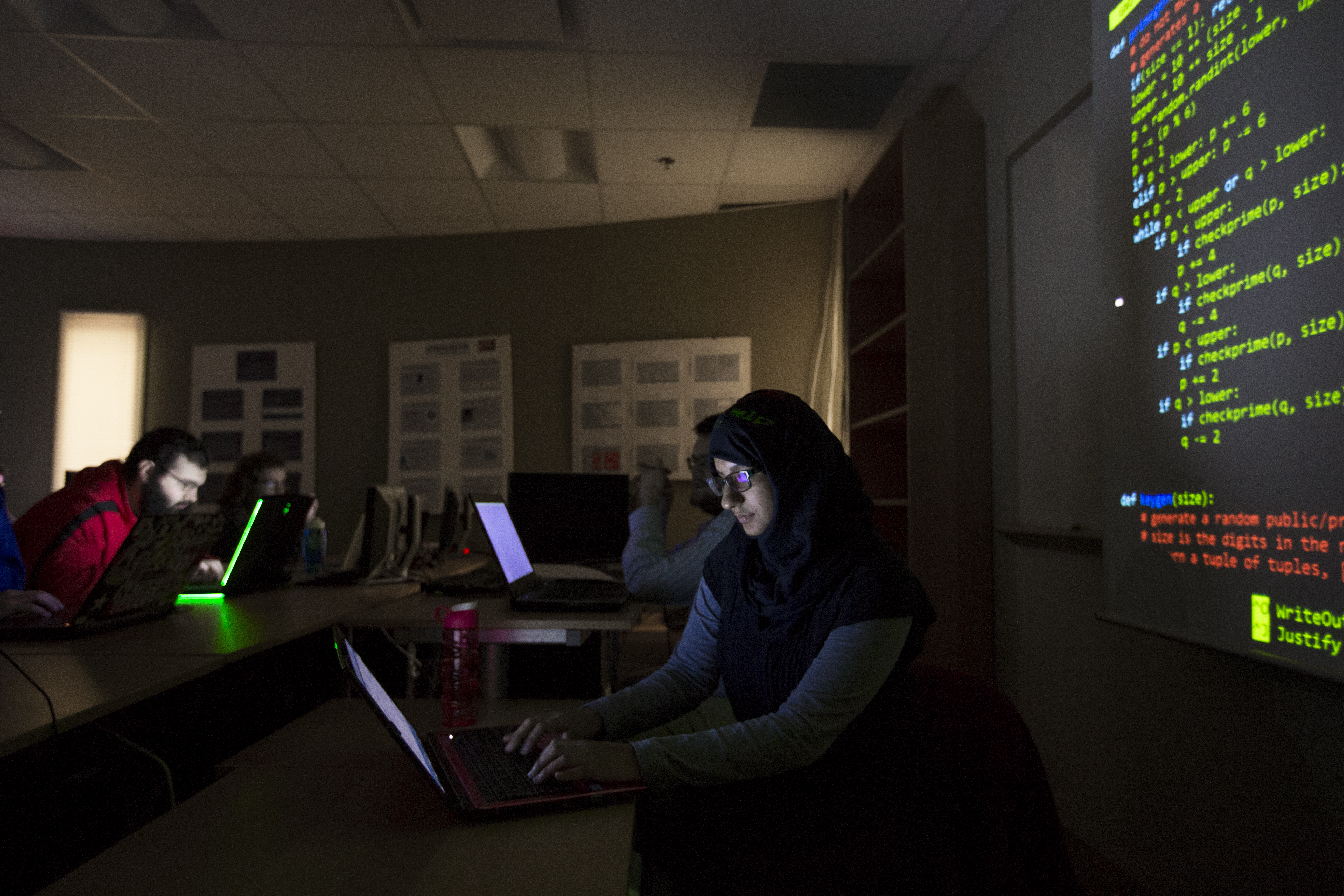 Student on laptop in dark lab with code projected on wall.