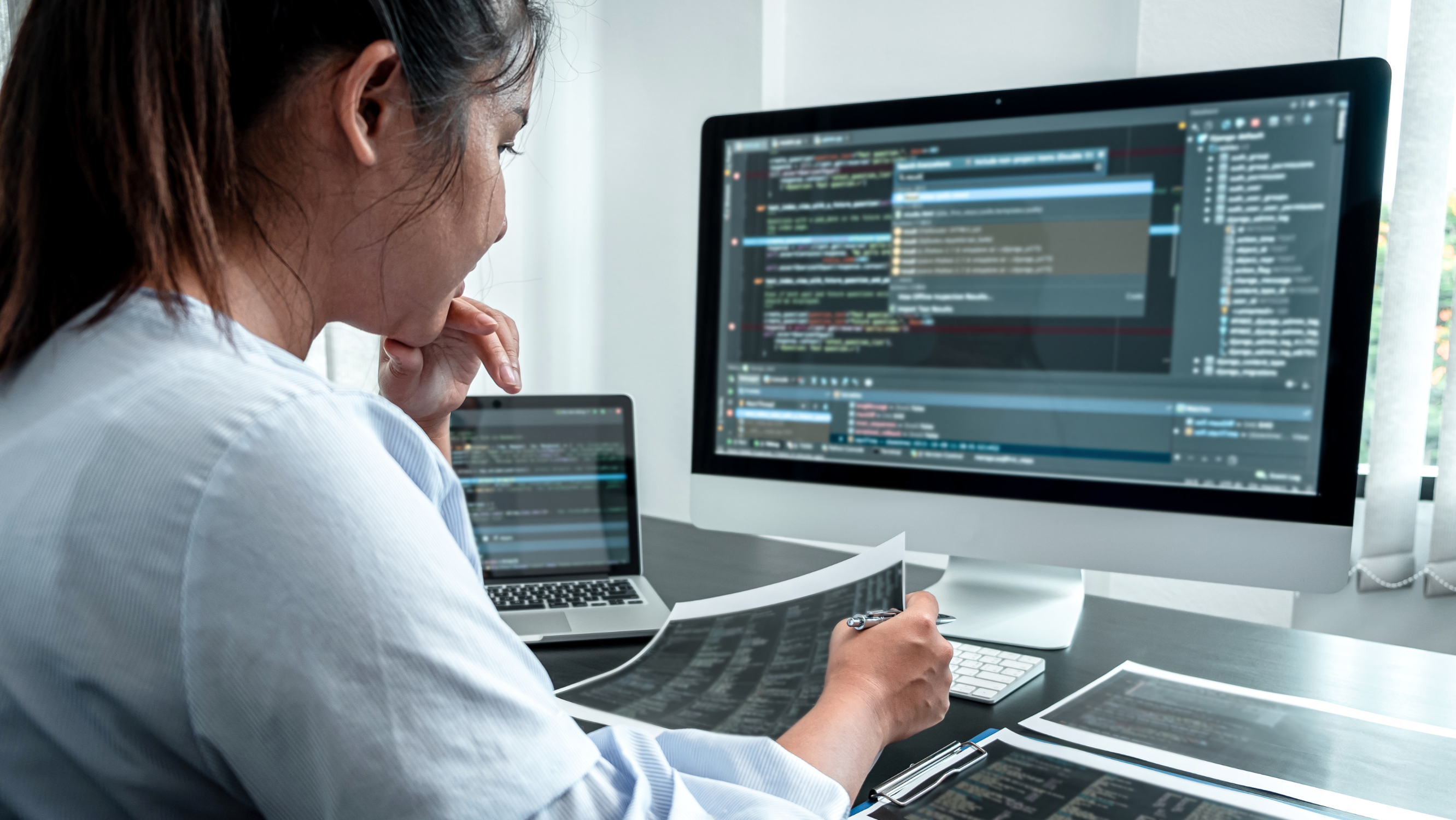 A woman in the foreground wearing a light blue and white striped shirt reviews code on two computer screens and multiple print-outs.