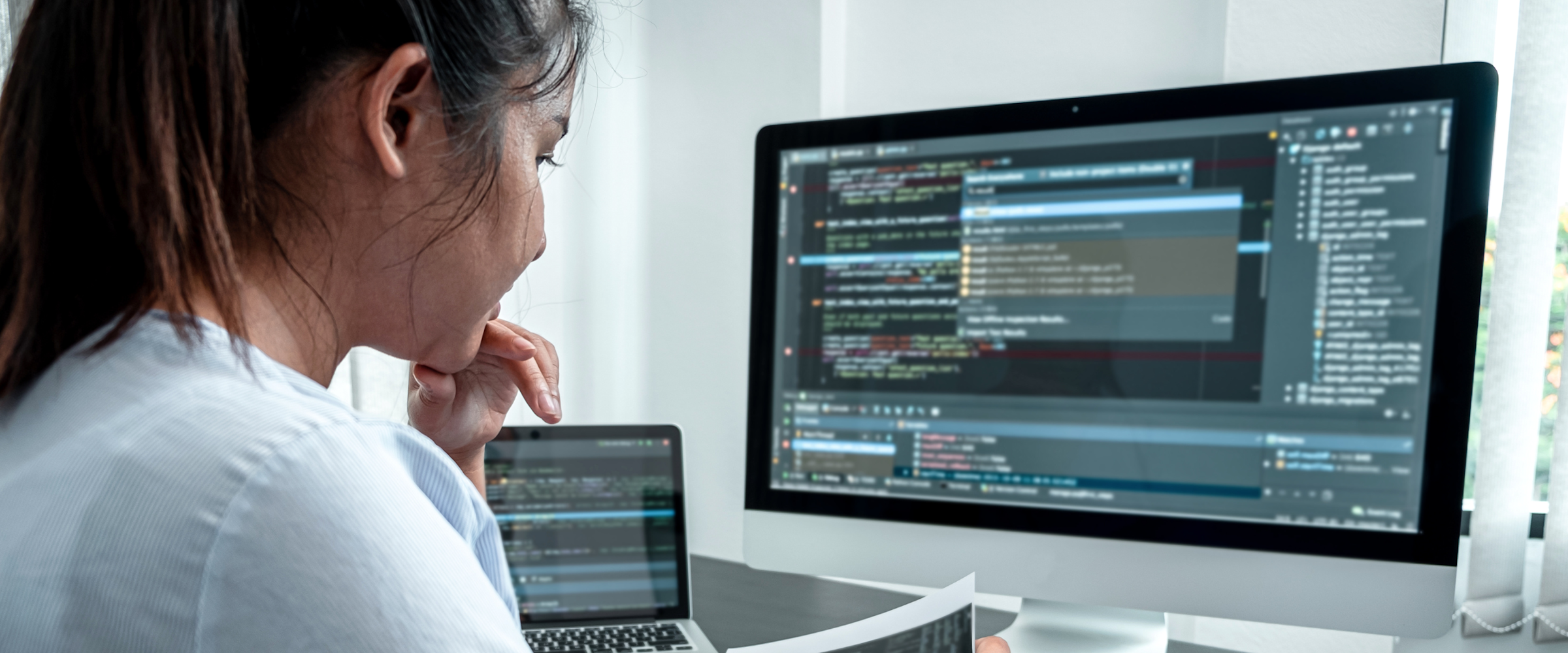 A woman in the foreground wearing a light blue and white striped shirt reviews code on two computer screens and multiple print-outs.