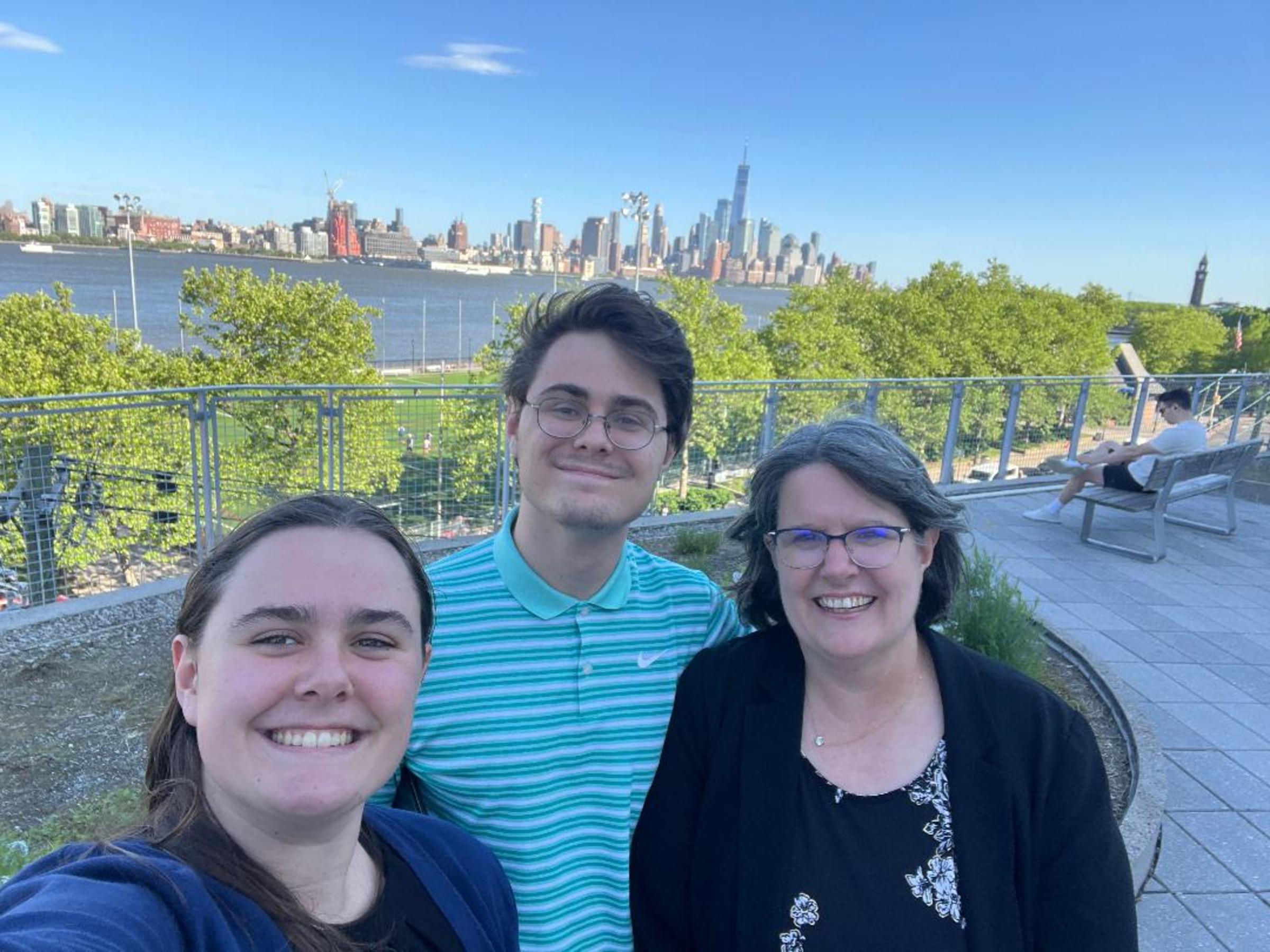 A photo of Patricia Muisener standing with her two children outdoors, with the Manhattan skyline and Hudson River in the background.