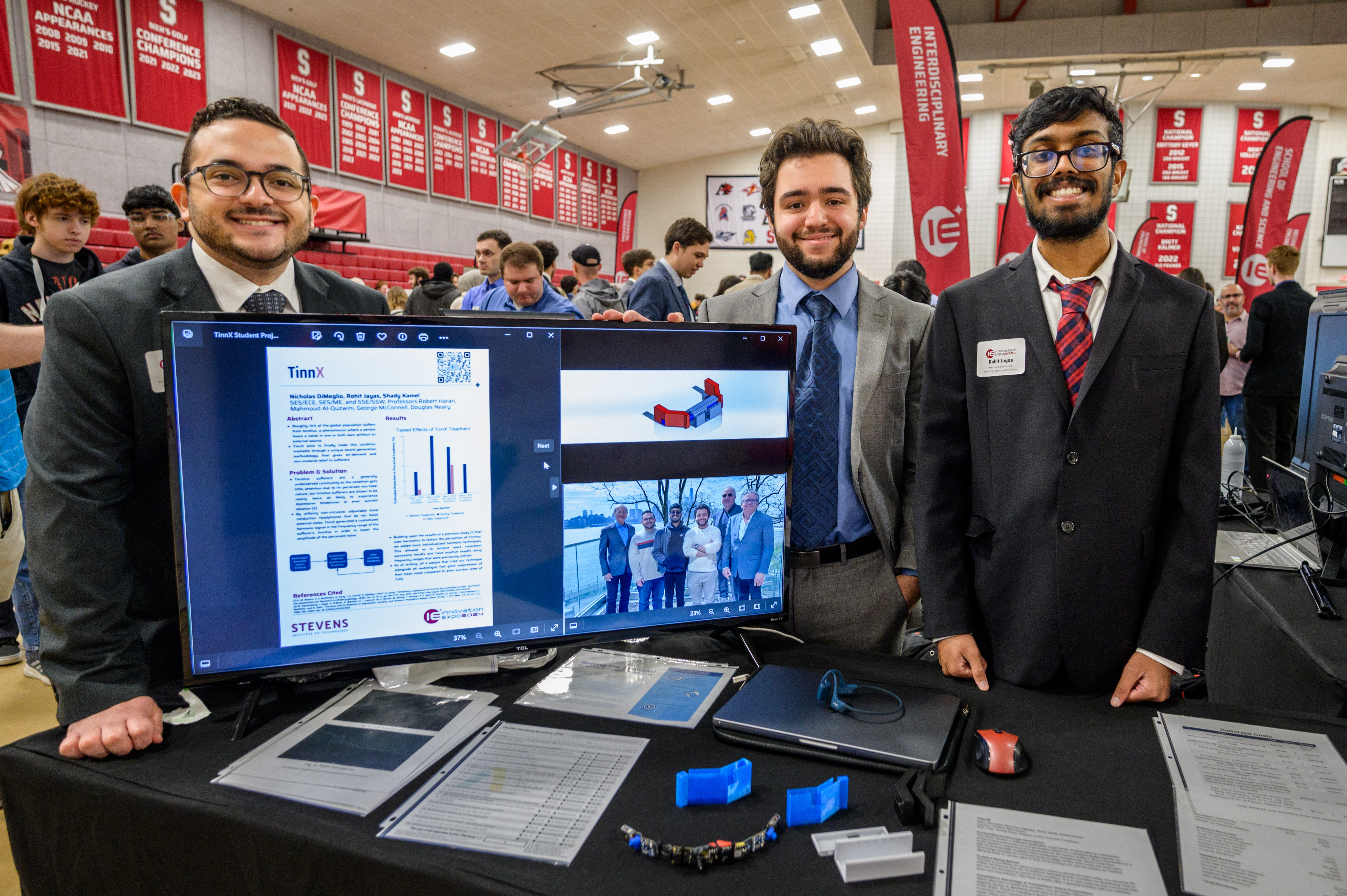 Three male students in a an exhibition hall with a computer screen displaying an abstract of their project work.