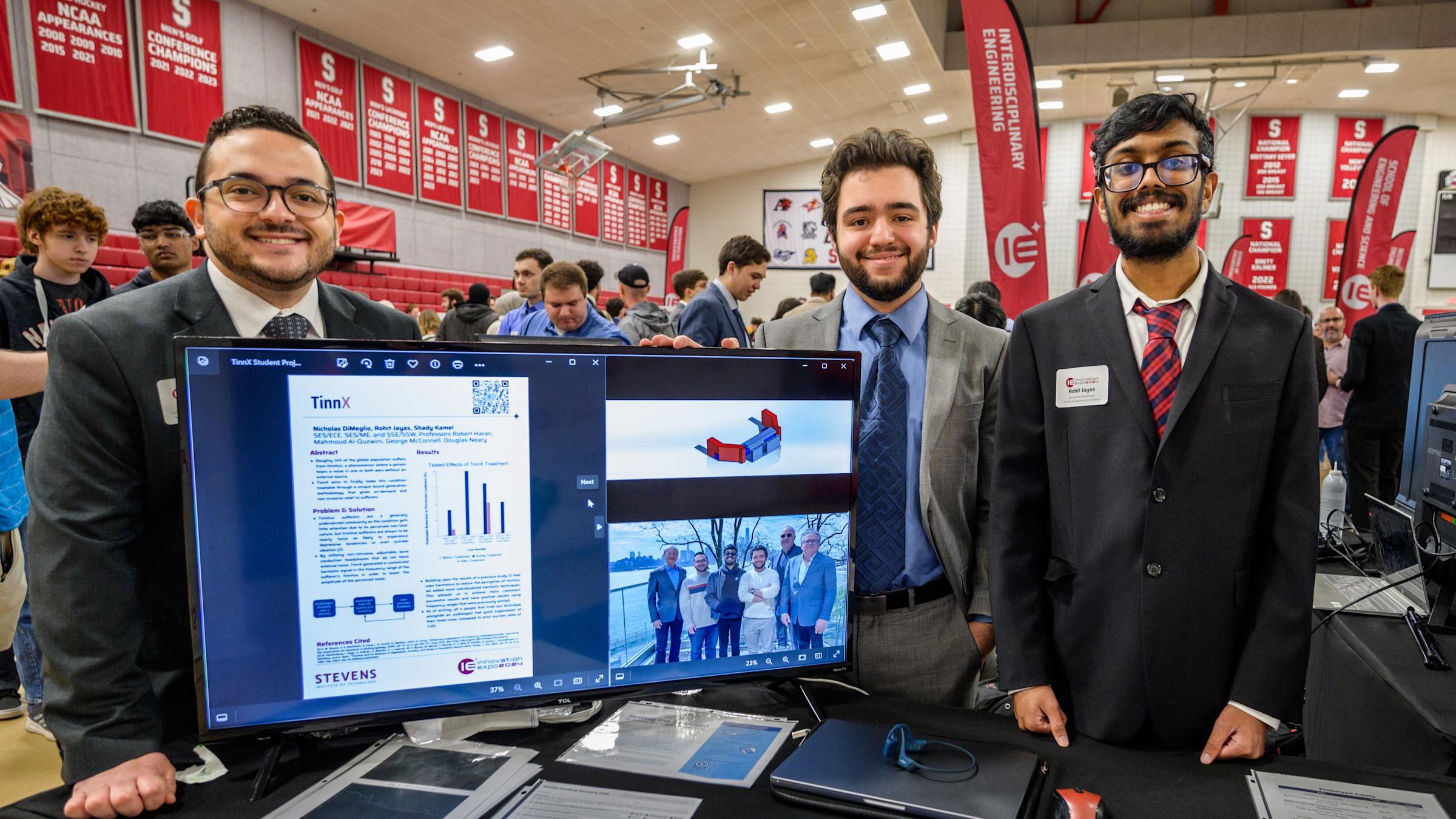 Three male students in a an exhibition hall with a computer screen displaying an abstract of their project work.