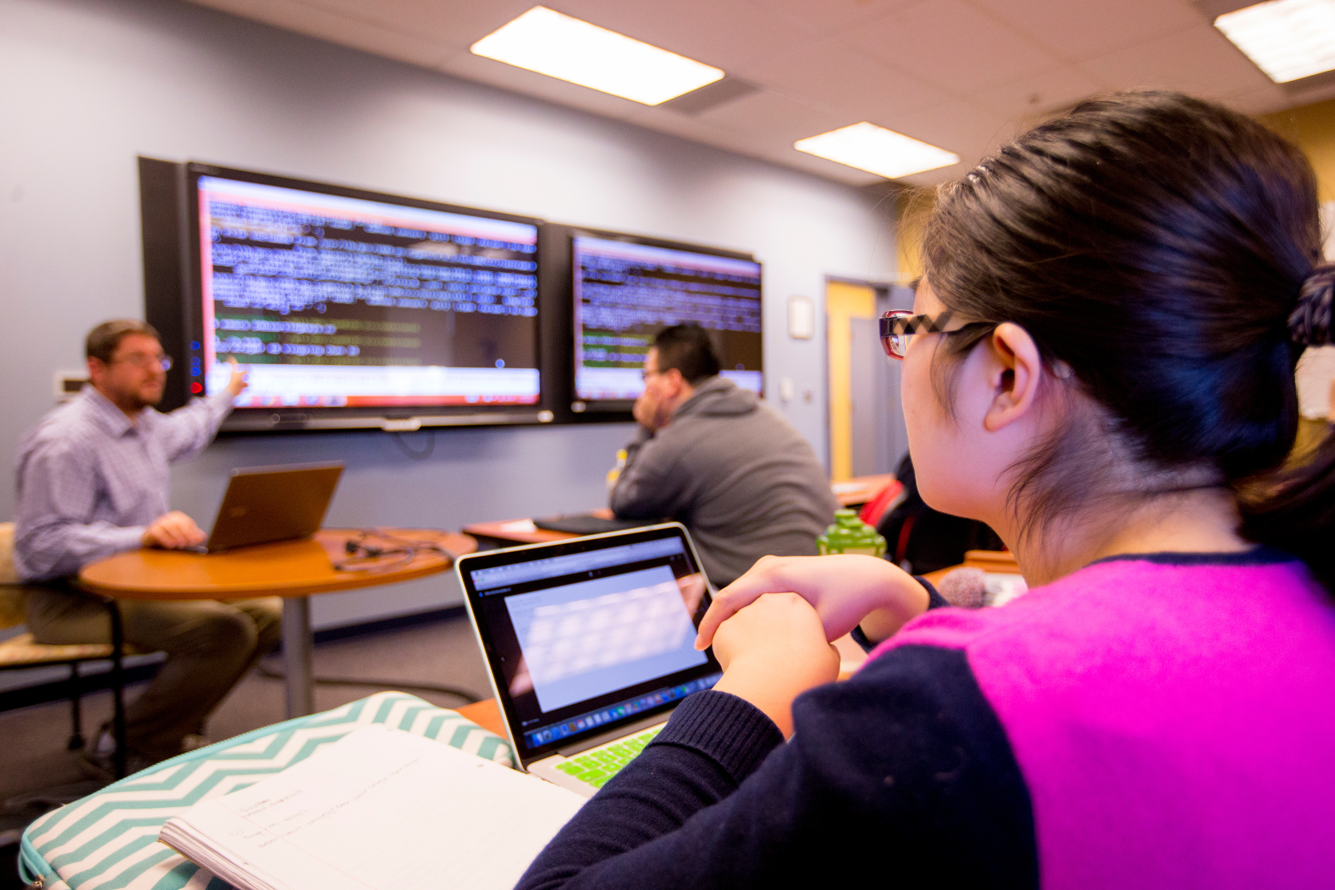 A student is seated with a laptop in the right side foreground watching a faculty member conduct a class using wall monitors.