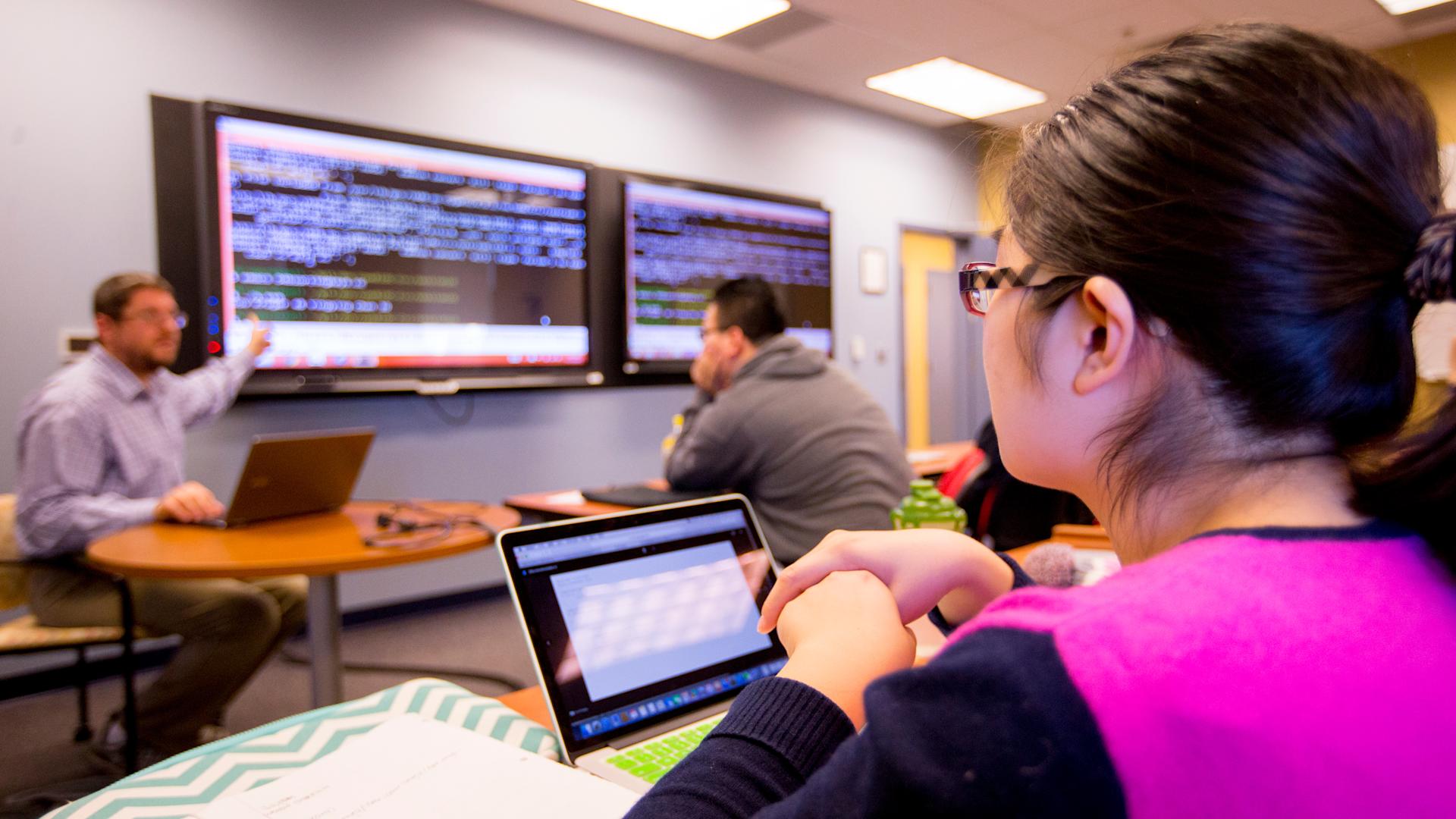 A student is seated with a laptop in the right side foreground watching a faculty member conduct a class using wall monitors.