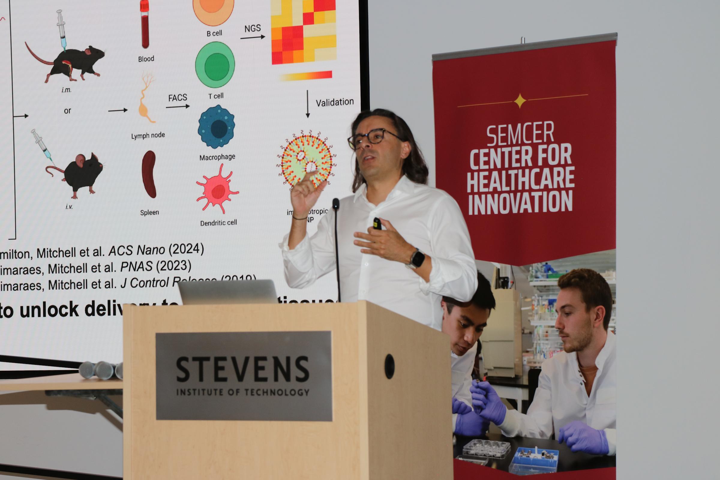Michael J. Mitchell, Associate Professor Bioengineering at the University of Pennsylvania, speaks from a podium in front of a large display.