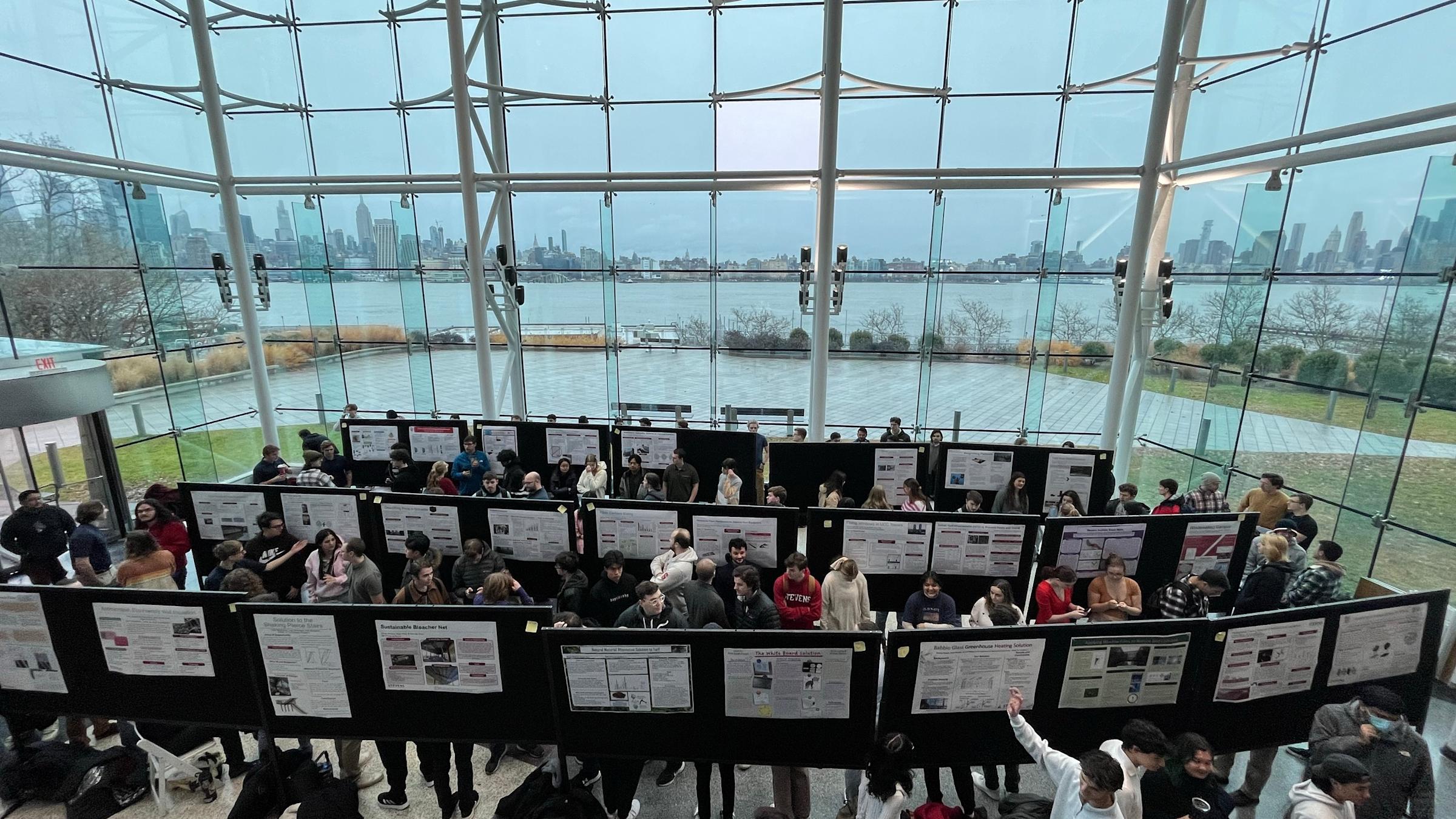 Students in a glass atrium area look at poster presentations.