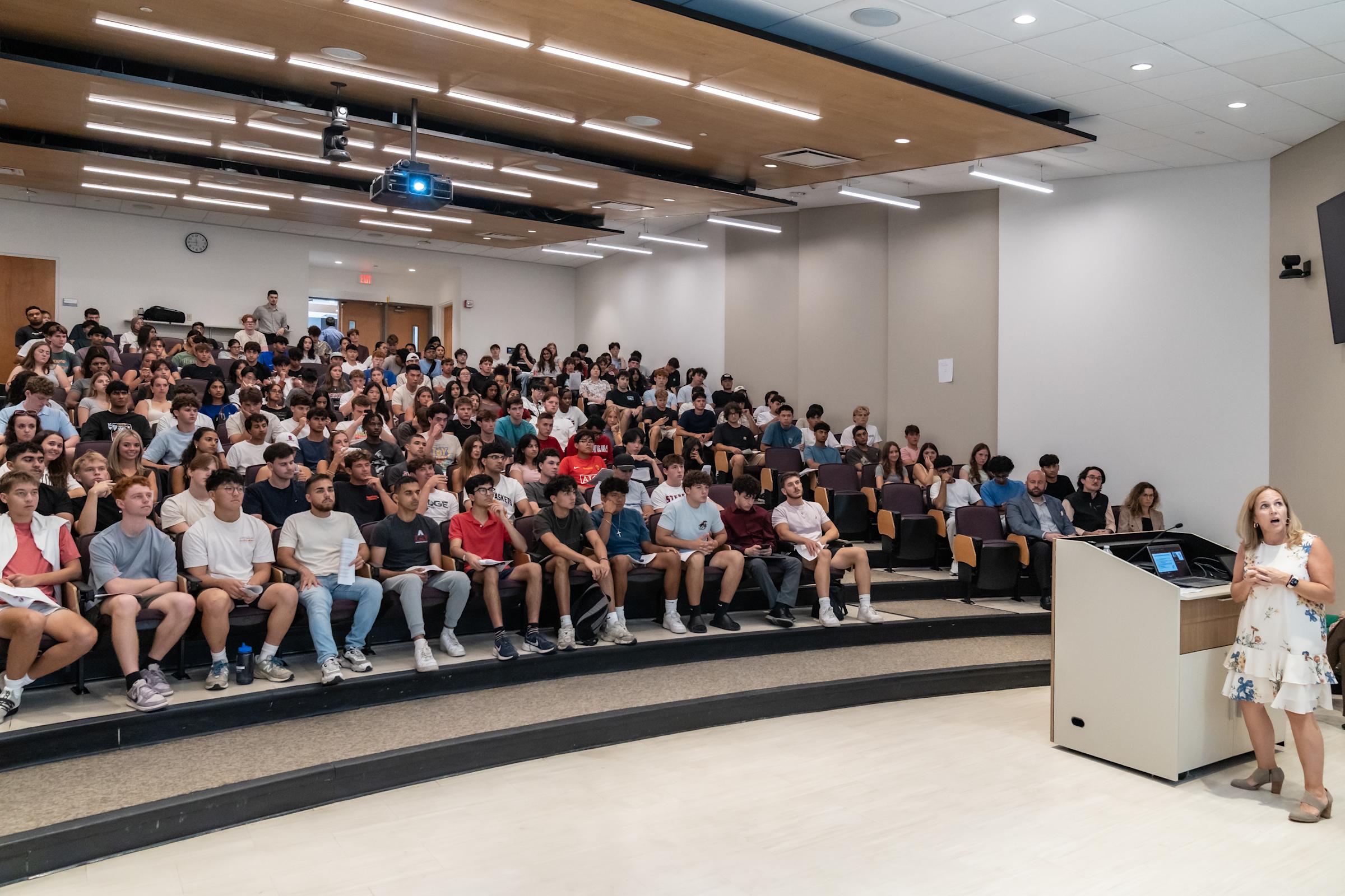 Blonde woman in floral dress presenting to crowded tiered lecture hall filled with college students in modern auditorium with wood accent ceiling.