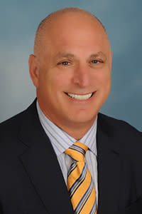 Professional headshot of a smiling man with a shaved head, wearing a dark suit, striped shirt, and yellow-and-blue striped tie, against a blue background.