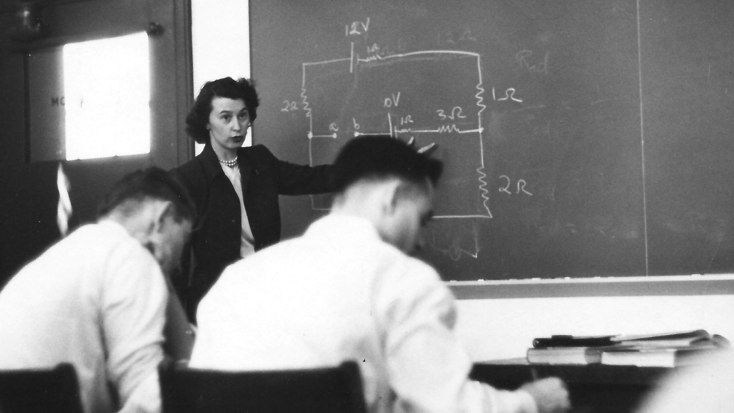 young woman from the 1940s standing at a chalk board speaking in front of a class, with a mathematical problem on the chalk board.