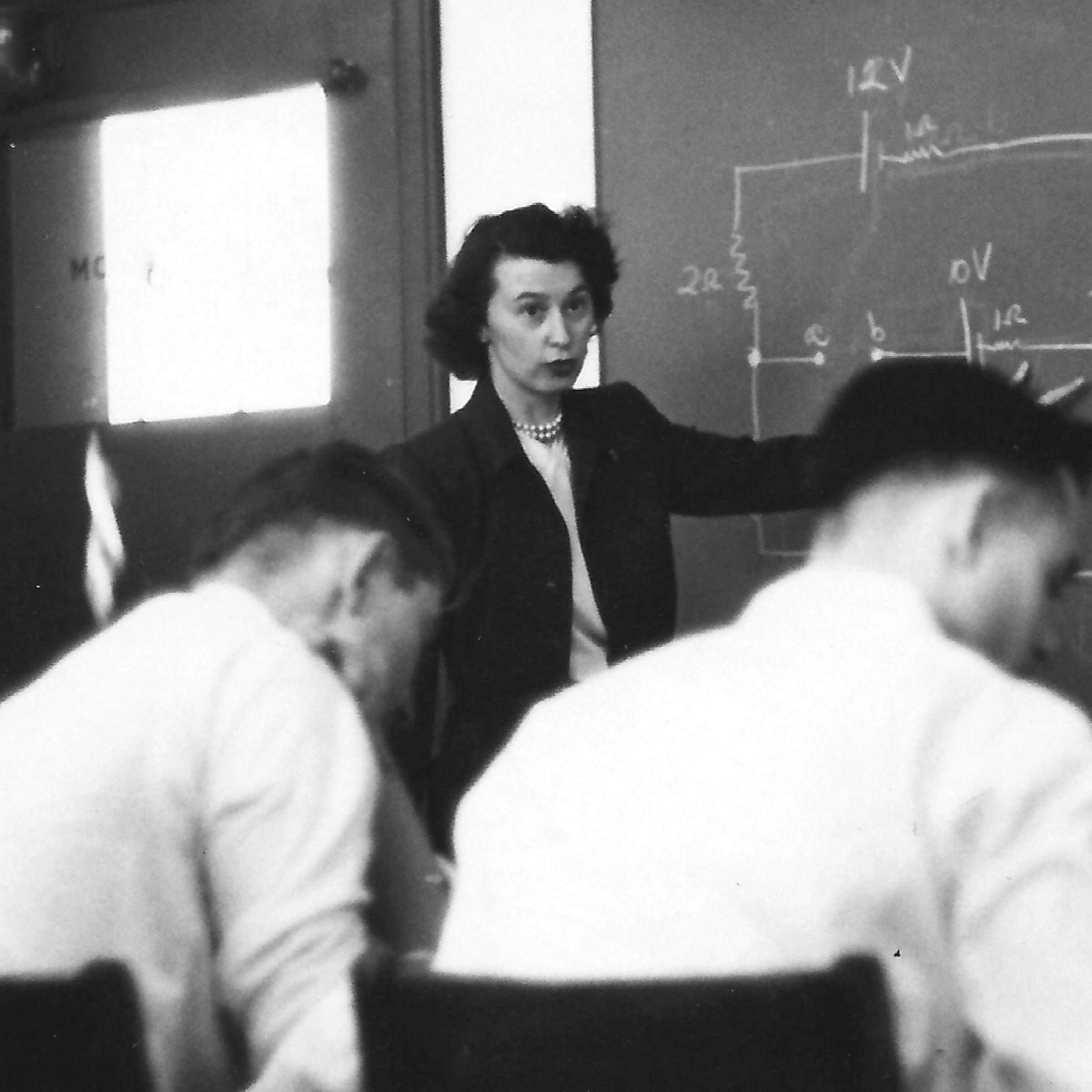 young woman from the 1940s standing at a chalk board speaking in front of a class, with a mathematical problem on the chalk board.