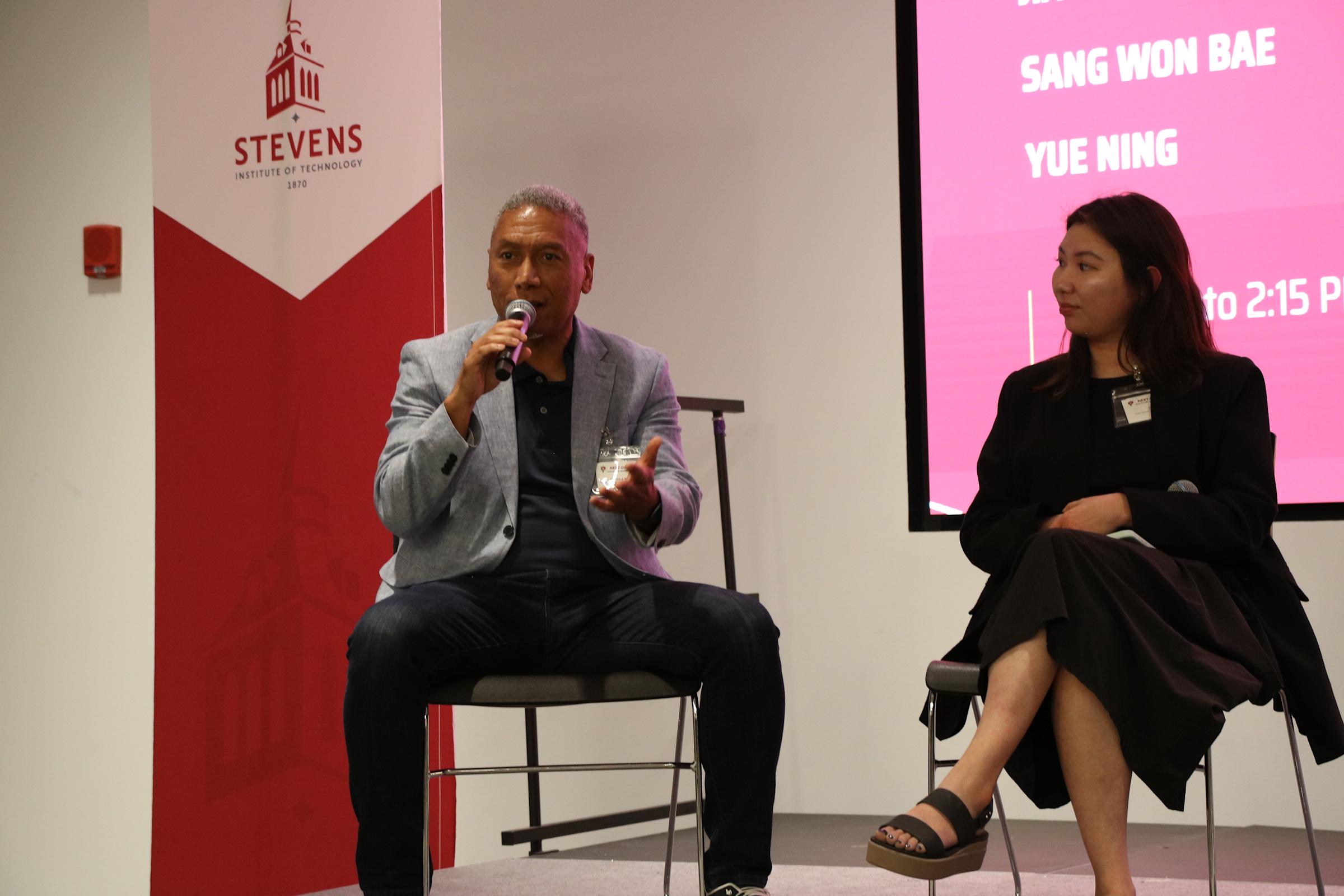 Lyndon J. Mitnaul speaking onstage into a microphone alongside Jia-Min Chu.