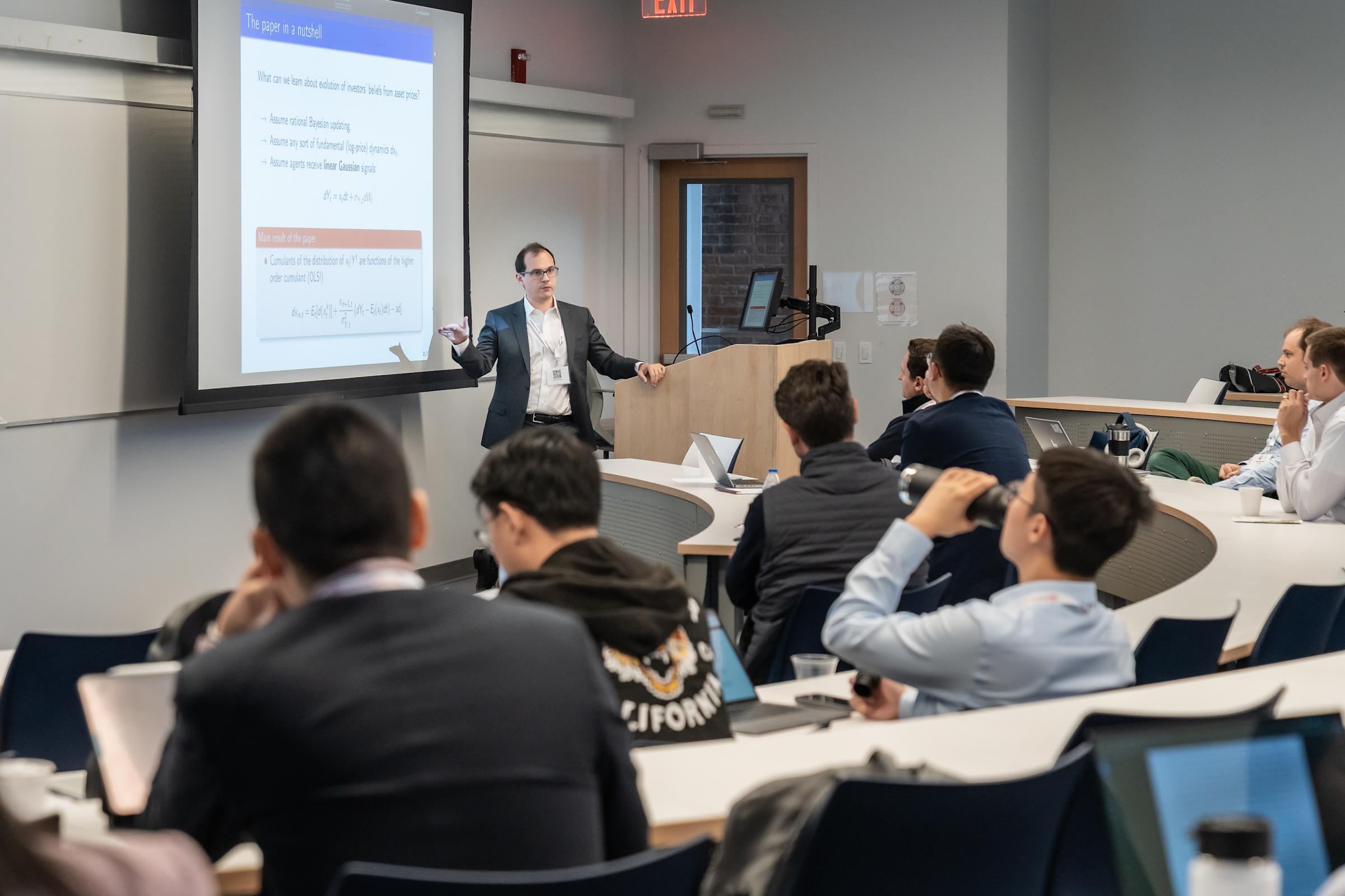 A presenter in glasses and suit jacket speaks to students in a lecture hall, gesturing toward a projected slide. Attendees sit at curved tiered desks with laptops, listening to the academic presentation in the modern classroom.