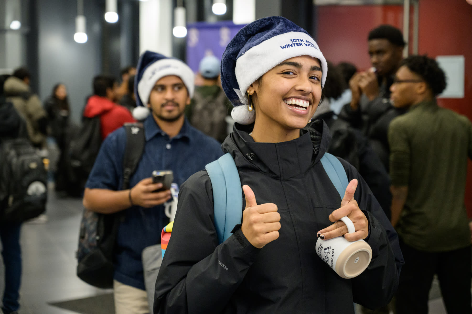 A student smiling and wearing a blue Santa hat that reads "10th Annual Winter Wonderland." The student is giving a thumbs up, showing excitement and enthusiasm.