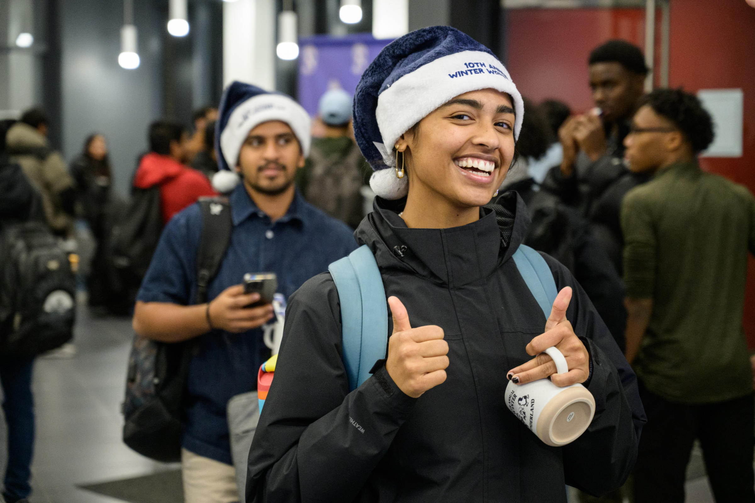 A student smiling and wearing a blue Santa hat that reads "10th Annual Winter Wonderland." The student is giving a thumbs up, showing excitement and enthusiasm.