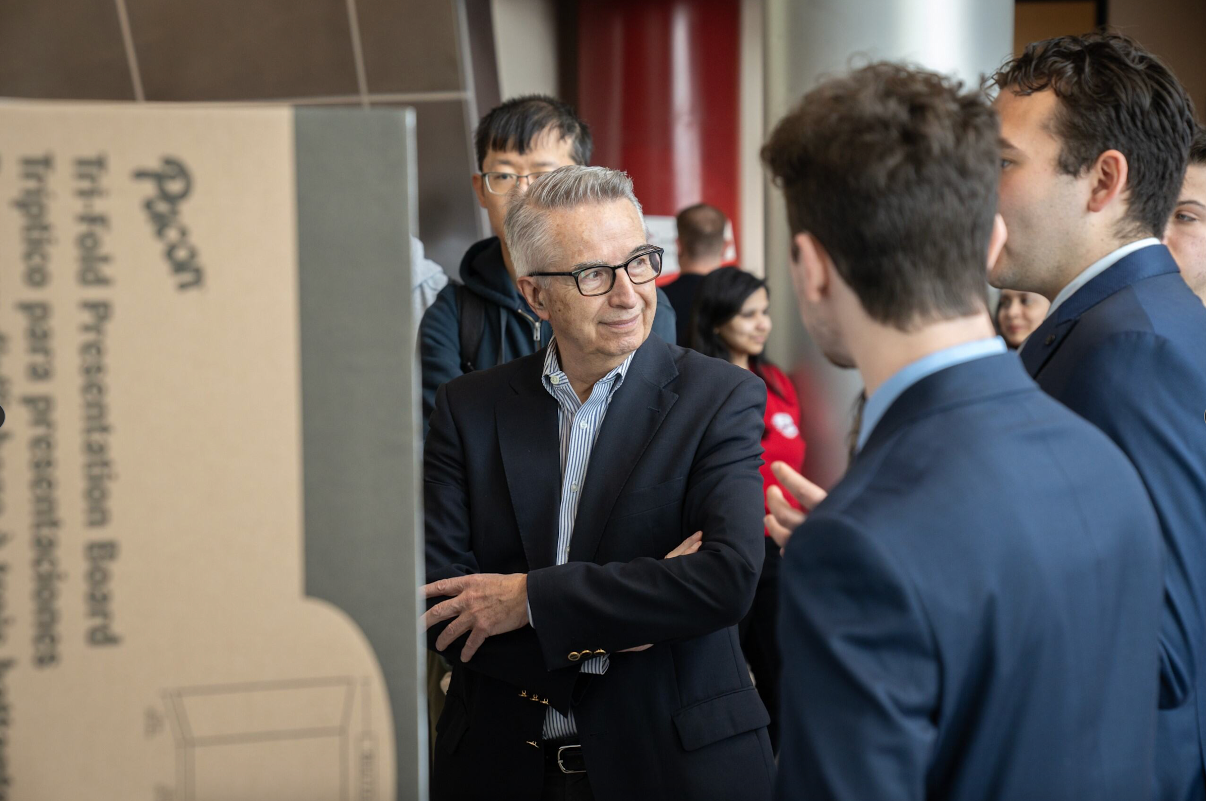 Gregory Prastacos, wearing a navy blue blazer, speaks with members of a research project team in front of their posterboard.