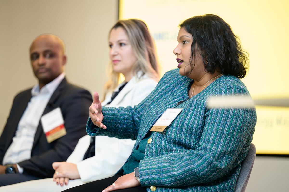 A panelist in a textured blue-green jacket speaks and gestures. Two fellow panelists are seated nearby, listening onstage as part of a professional networking event.