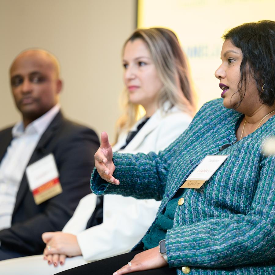 A panelist in a textured blue-green jacket speaks and gestures. Two fellow panelists are seated nearby, listening onstage as part of a professional networking event.