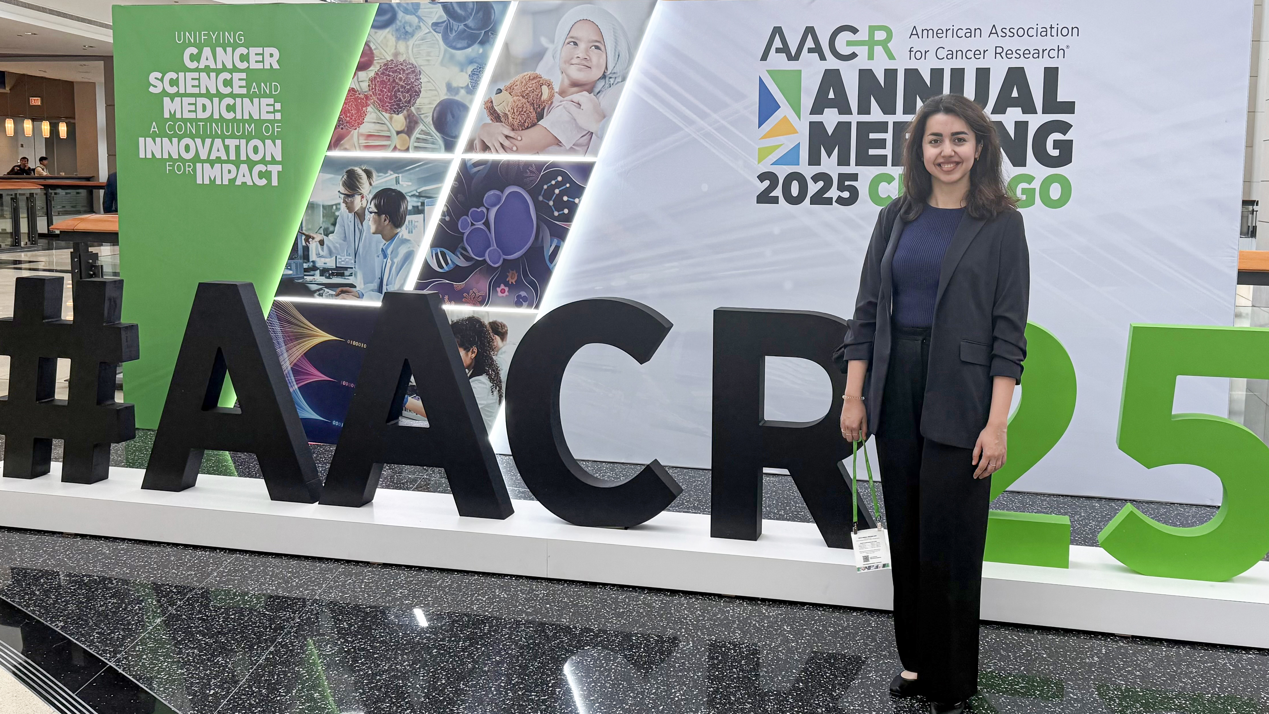 A young woman in business attire stands in front of a convention sign with the hashtag AACR25 in front of it.