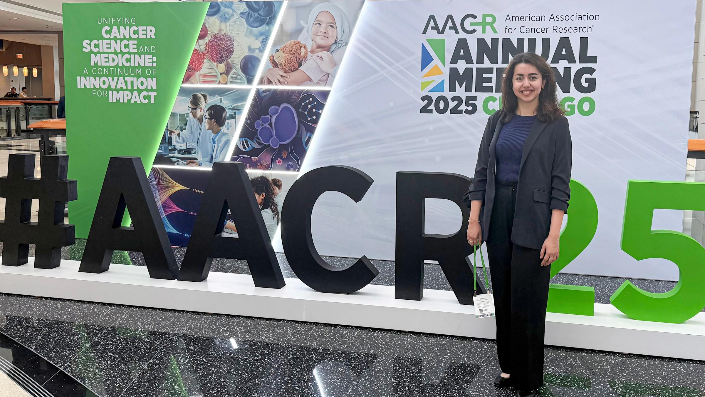 A young woman in business attire stands in front of a convention sign with the hashtag AACR25 in front of it.