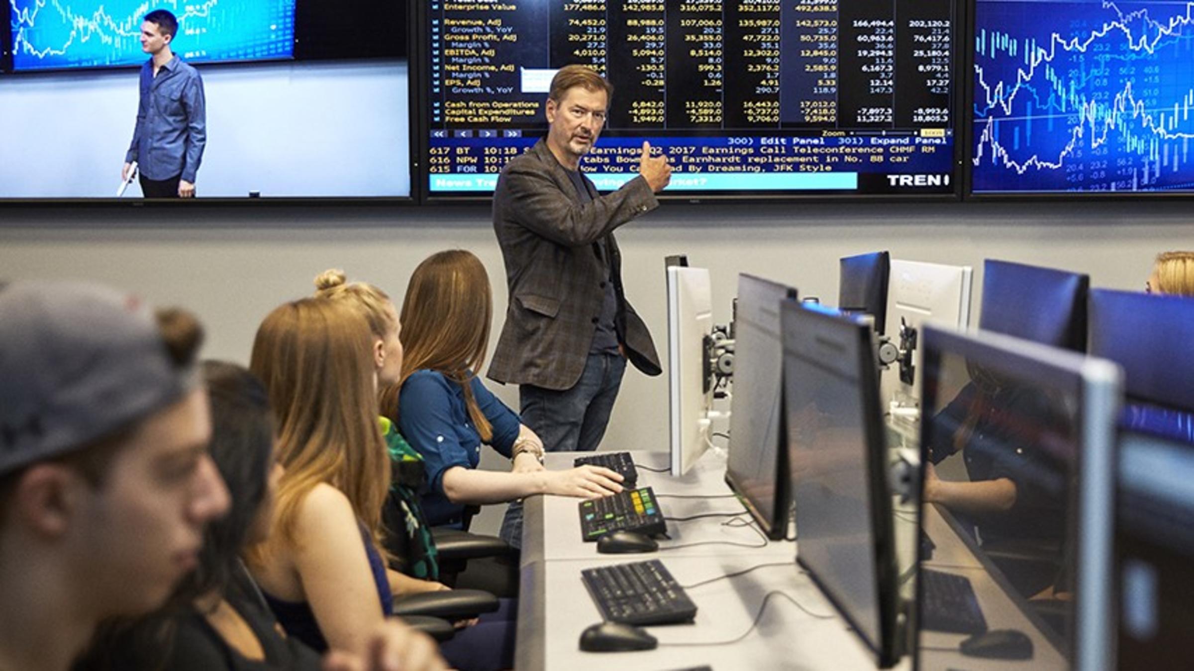 George Calhoun, in a checked jacket, gestures to Bloomberg data on a large screen as he teaches a class in the Hanlon lab.