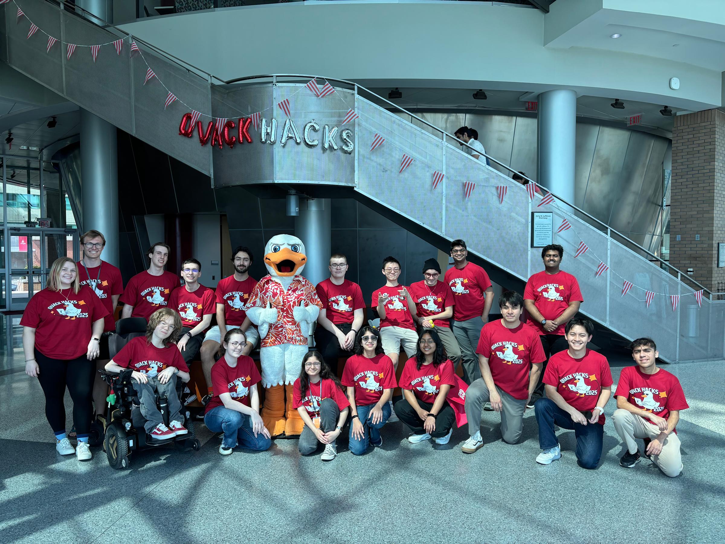 A group of students smile at the camera in two rows. The back row is standing and the front row is sitting. Atilla the duck character is with the students. Balloons that say "QUACK HACKS" is on the wall behind them.