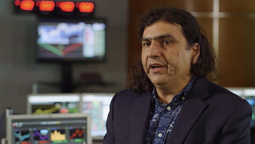 Ionut Florescu, in a dark shirt and jacket, speaking in the Hanlon Lab. Bloomberg terminals are visible in the background.