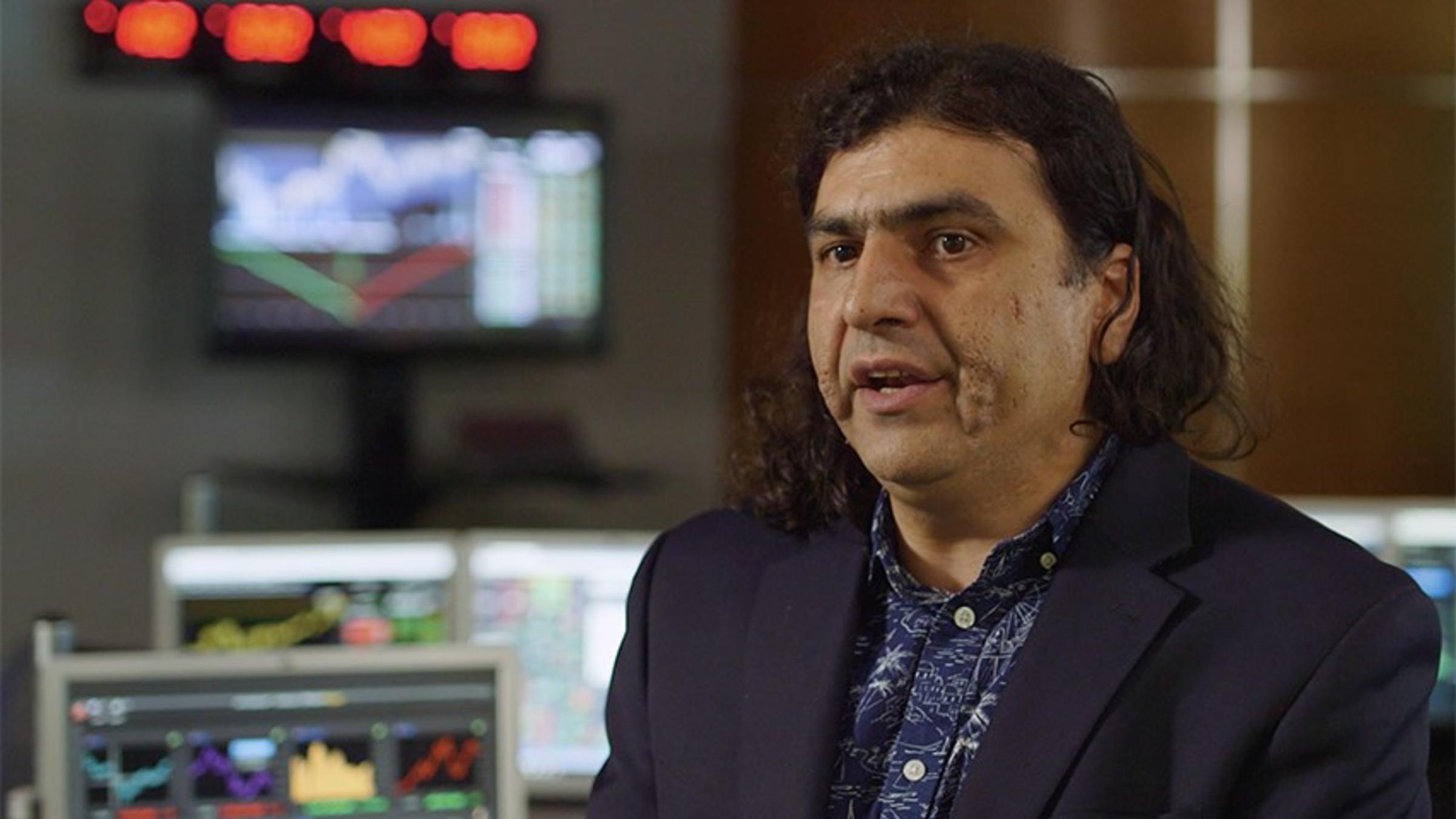 Ionut Florescu, in a dark shirt and jacket, speaking in the Hanlon Lab. Bloomberg terminals are visible in the background.