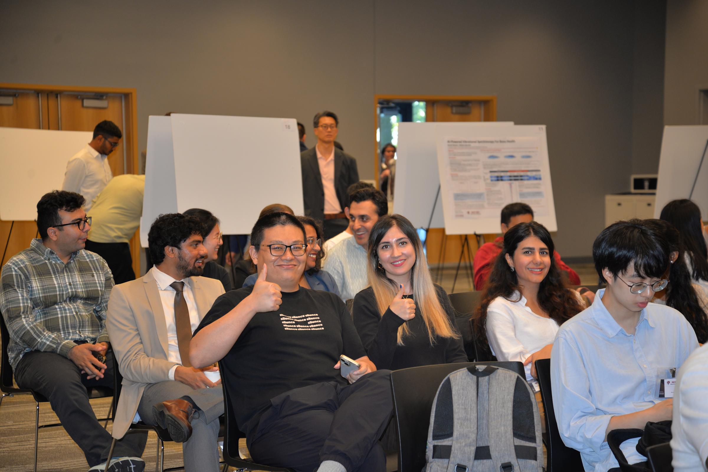 Two students, one giving a thumbs up sign, sit in an audience.