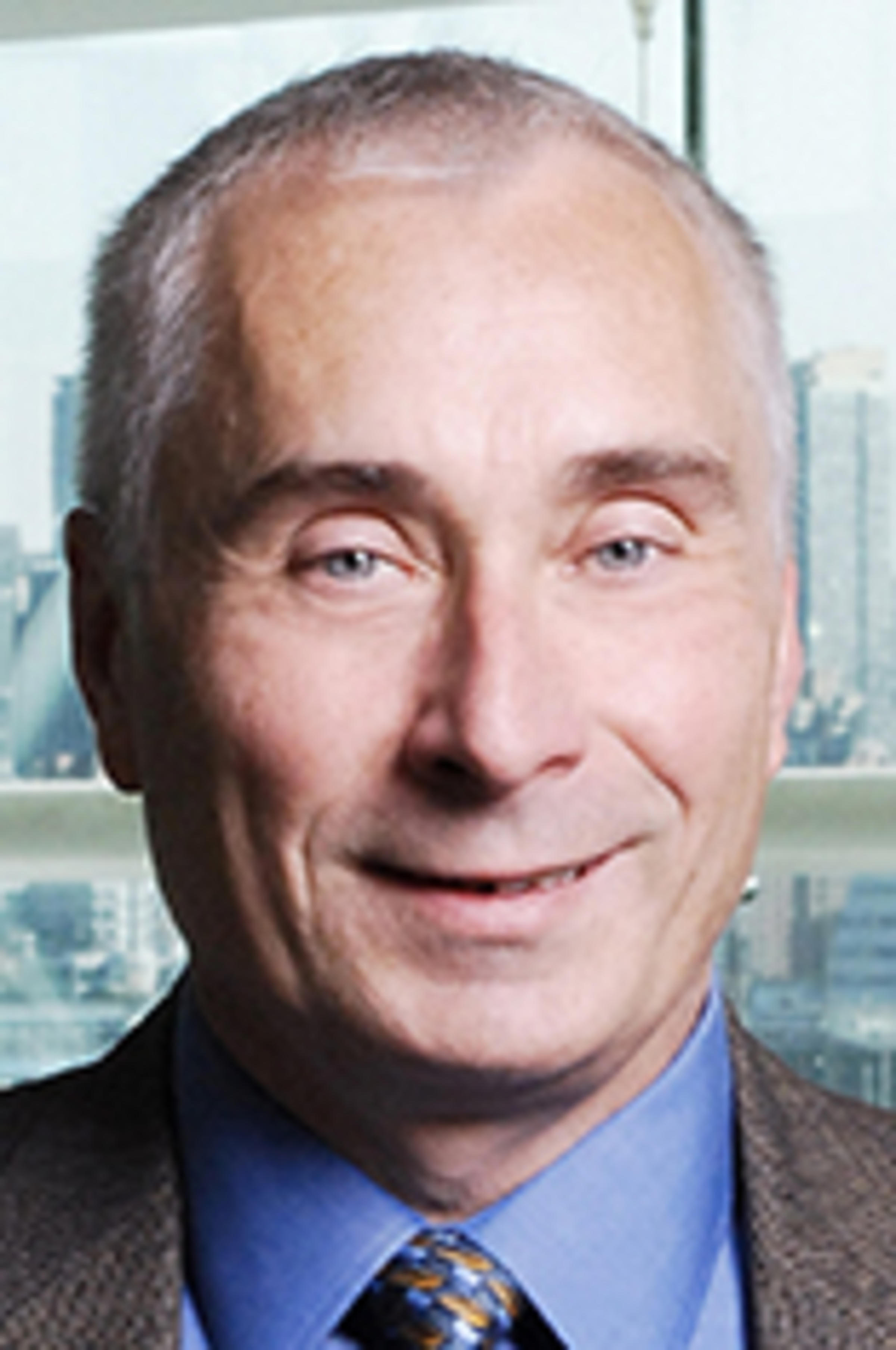 A headshot of Chris Asakiewicz, wearing a tan jacket and standing before the New York City skyline.