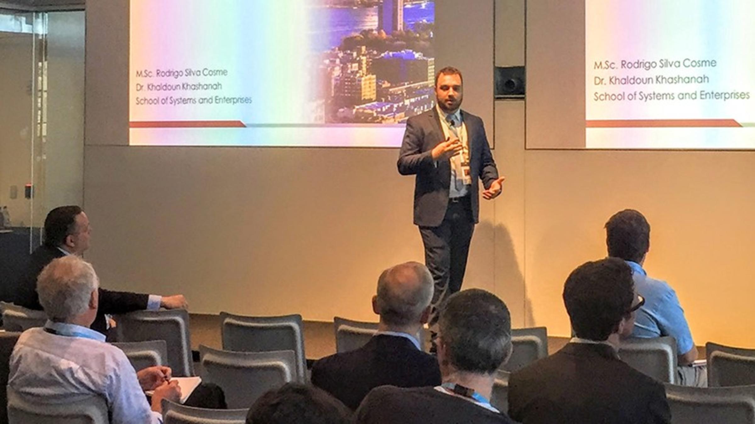 Rodrigo Silva Cosme leads a lecture in a crowded conference room. Two screens in the background show his Stevens thesis presentation.
