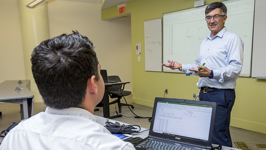 Alkis Vazacopoulos in front of a whiteboard in a classroom. A student in the foreground faces him.