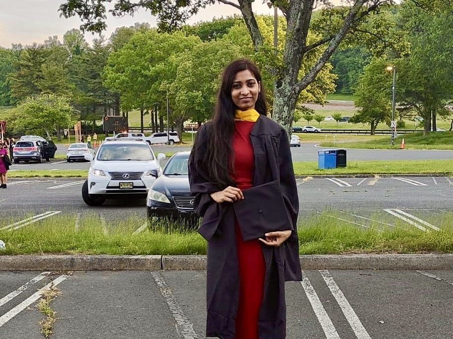 Student Shivani Bhawsar stands in a parking lot wearing her cap and gown for graduation