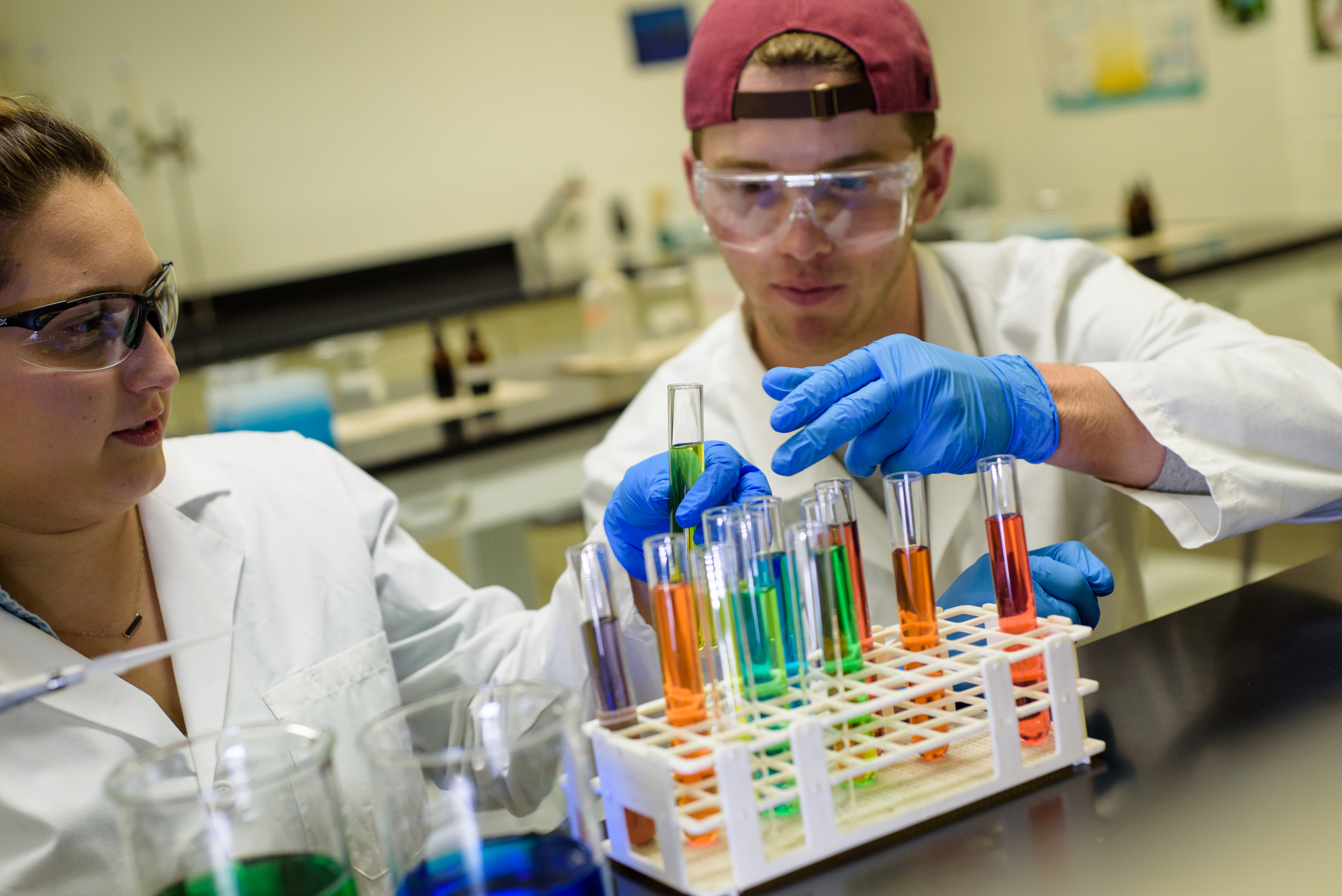 A female and male student with goggles in a lab examining liquids in colorful tubes