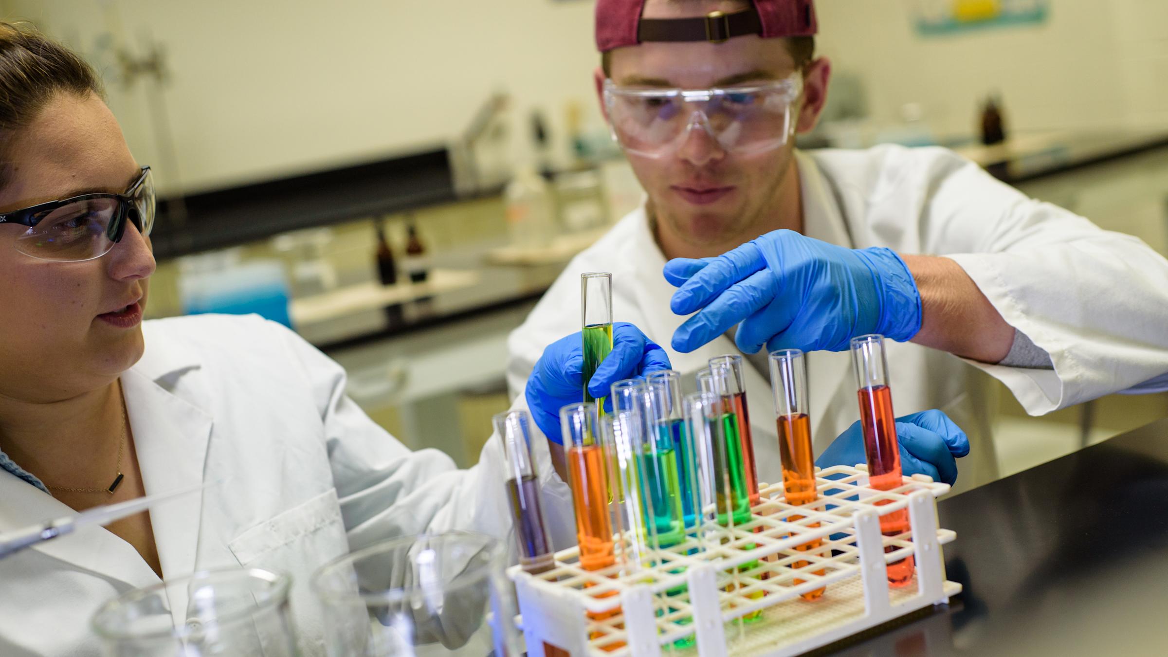 A female and male student with goggles in a lab examining liquids in colorful tubes