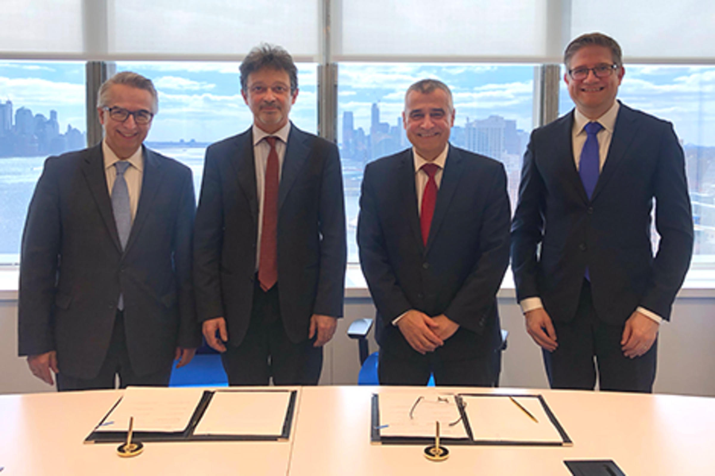 Four men stand behind a table with signed documents in front of them. New York City's skyline is visible in the back.