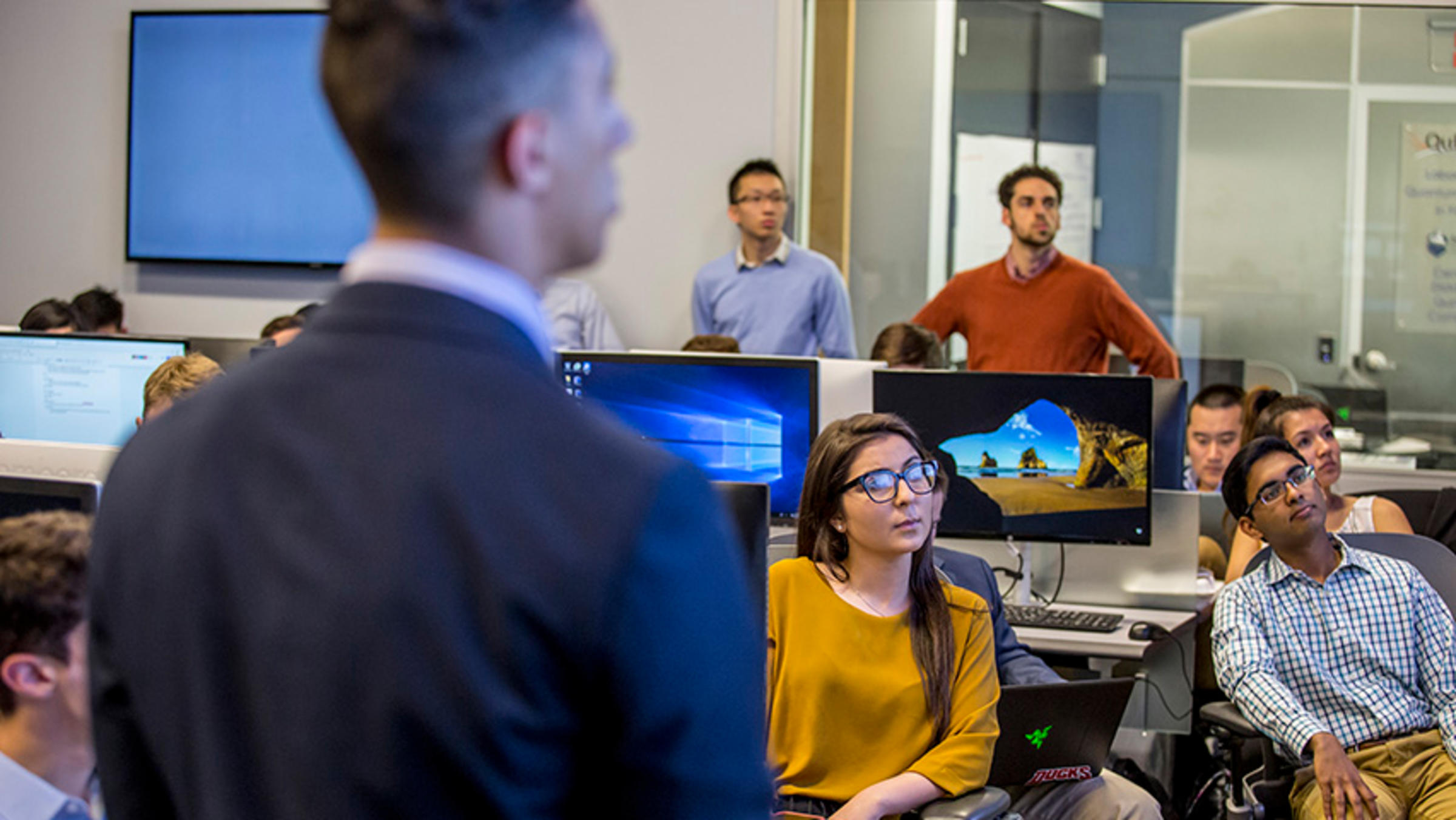 A group of students seated at computer terminals listens as another student leads a pitch in front of the classroom.