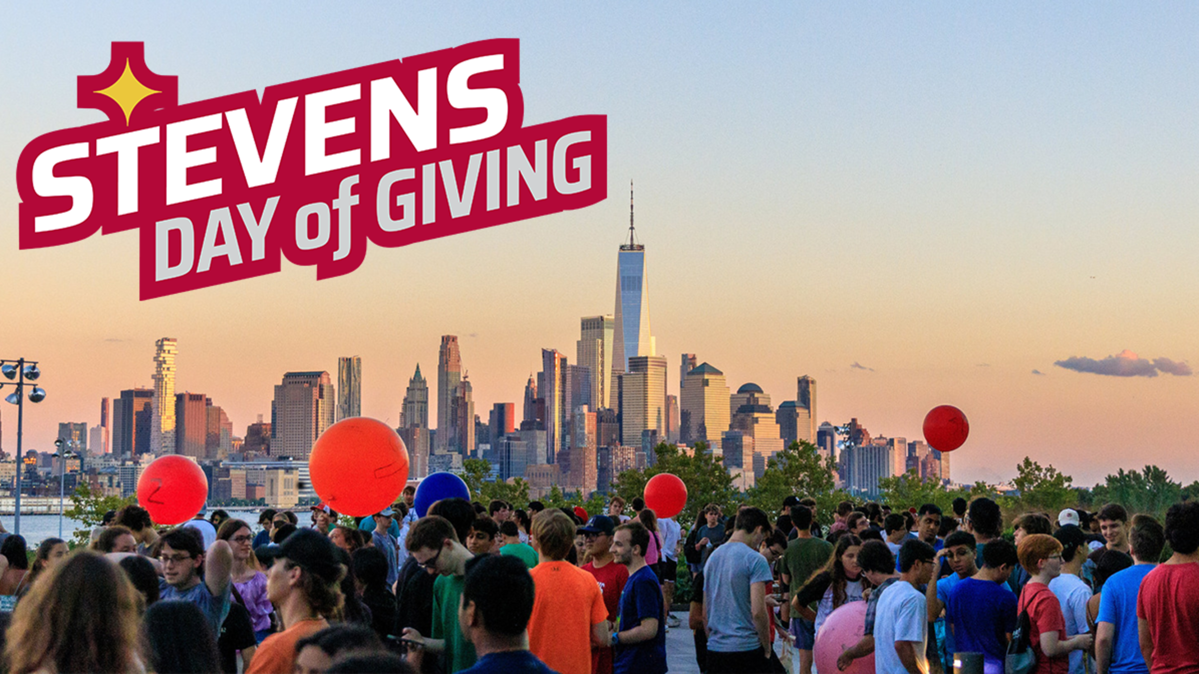Picture of students gathered on an outdoor patio. Large red balloons float amount the audience. In the background is part of the Southern New York City skyline on a cloudless evening.