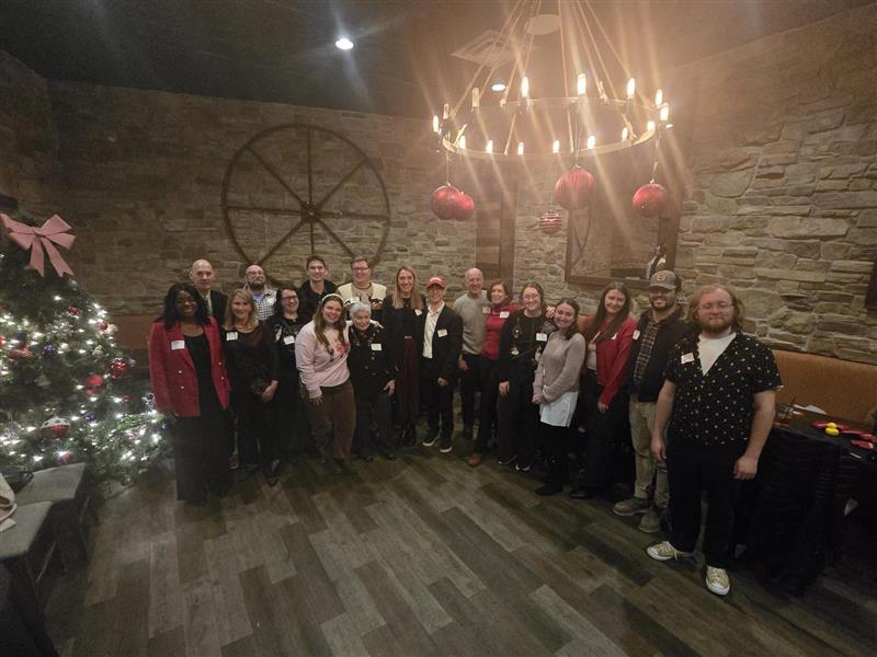 Attendees pose for a group photo at the Philadelphia Alumni Holiday Gathering 2025, standing beside a decorated holiday tree in a private event space with stone walls, festive ornaments and warm lighting.