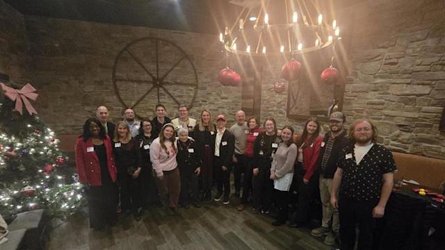 Attendees pose for a group photo at the Philadelphia Alumni Holiday Gathering 2025, standing beside a decorated holiday tree in a private event space with stone walls, festive ornaments and warm lighting.