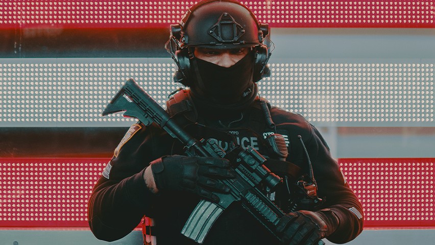 A police officer standing in front of red and white lights armed with an automatic weapon. CREDIT: Alec Favale, Unsplash
