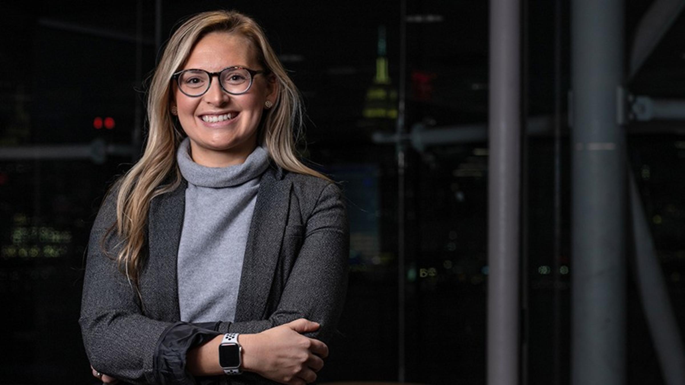 A young woman in a suit stands before the New York City skyline at night.