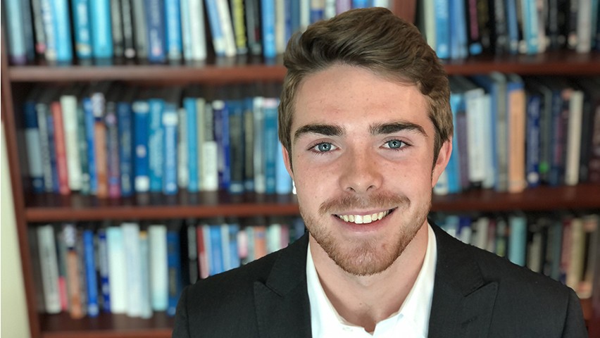Garrett Kincaid, in a dark jacket and dress shirt, stands in front of shelves of books at the School of Biusiness library.