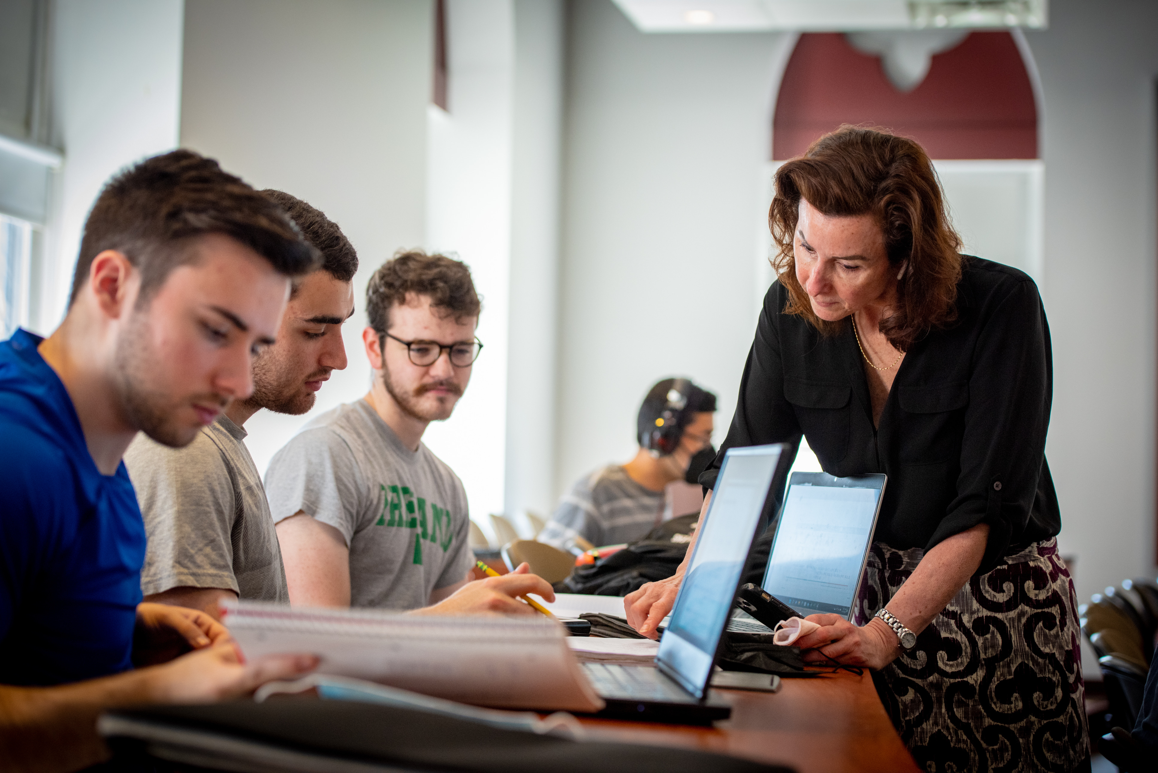 Four male students at a desk woking on their laptops while professor Kate Abel leans in to help