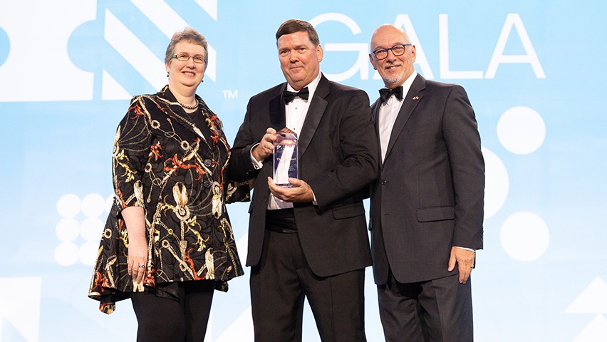 Three people in formal dress onstage. The PMI logo is visible on the bright blue background.
