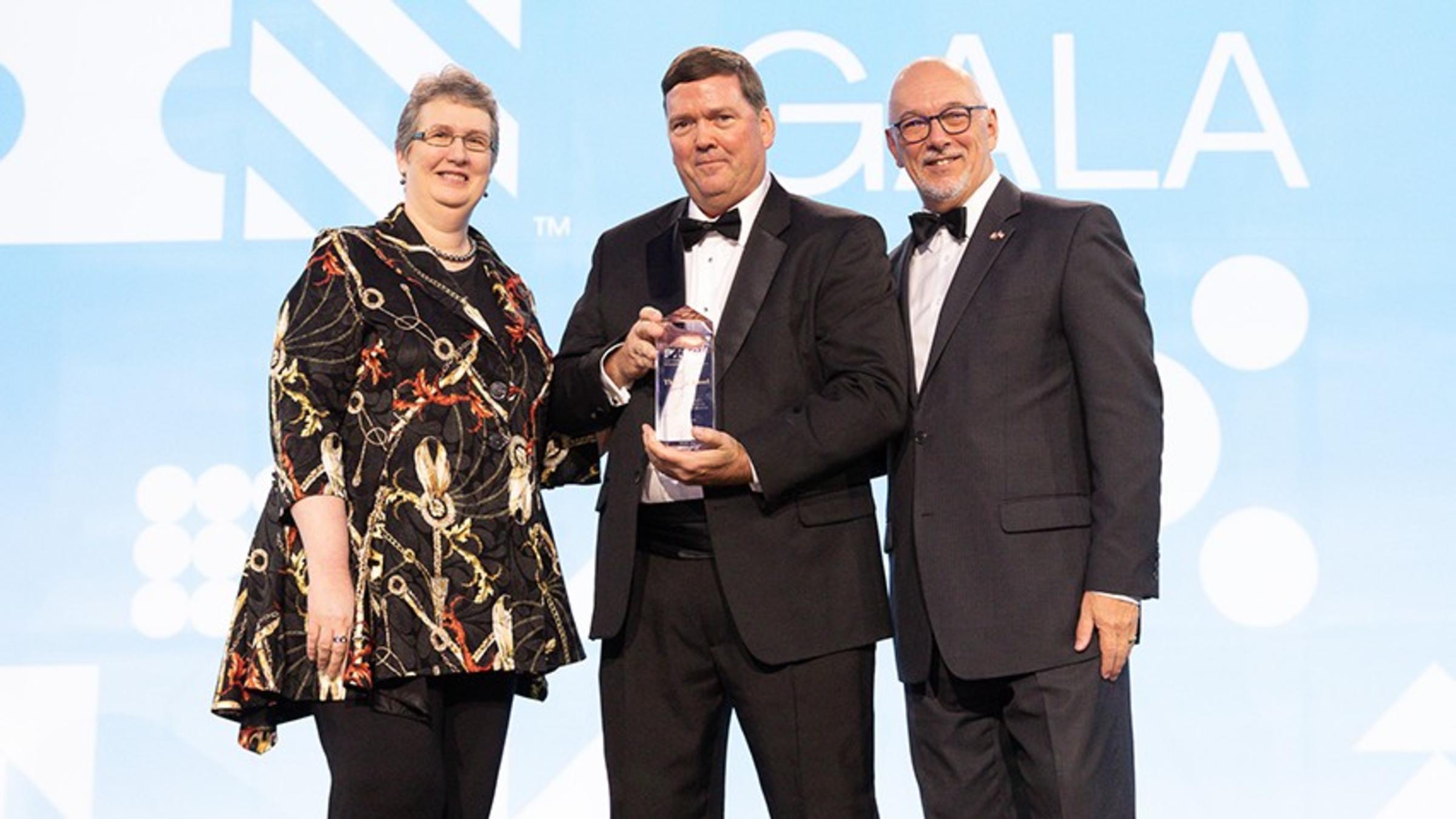 Three people in formal dress onstage. The PMI logo is visible on the bright blue background.