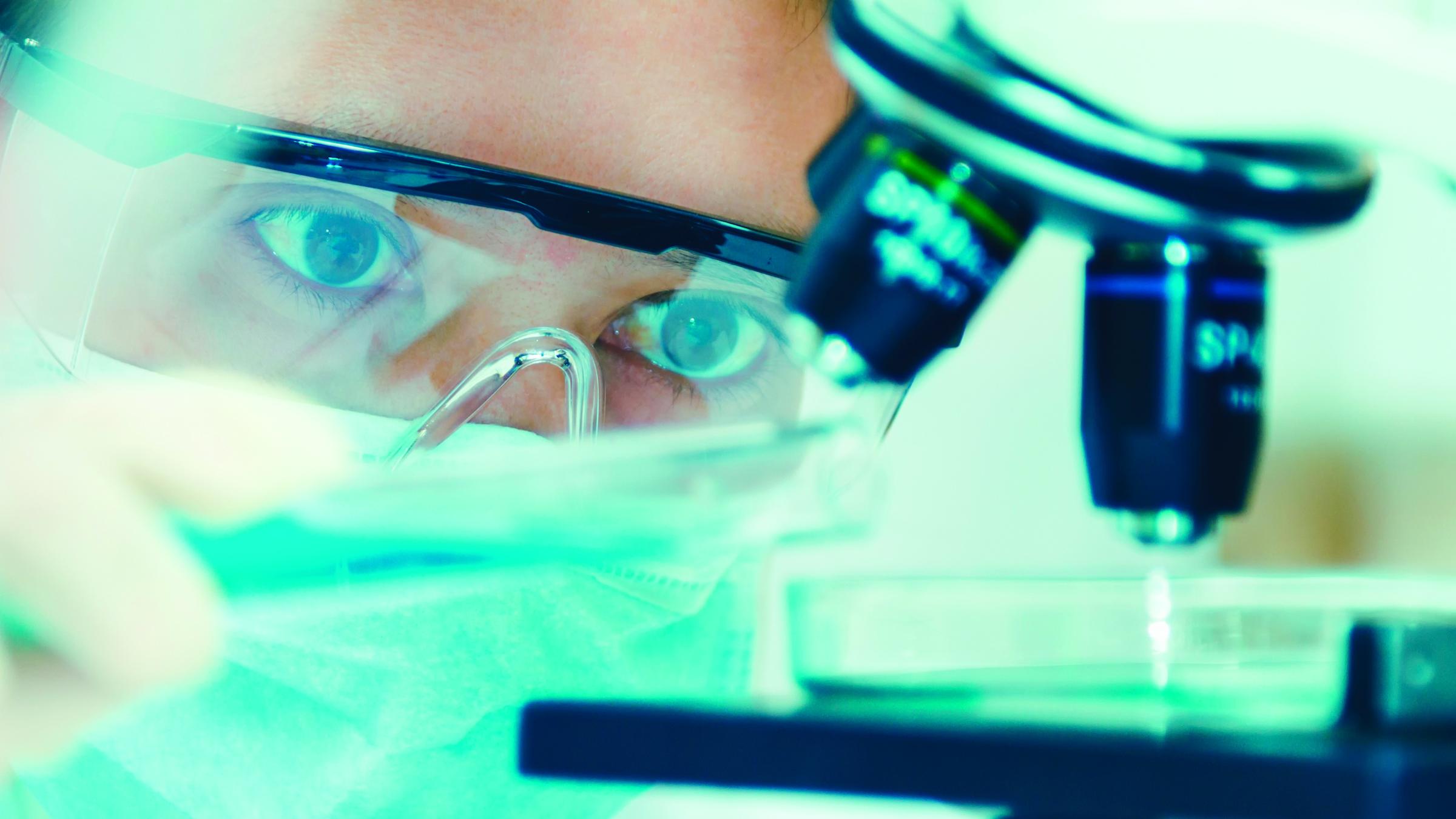 A woman in a clean suit views a sample under a microscope