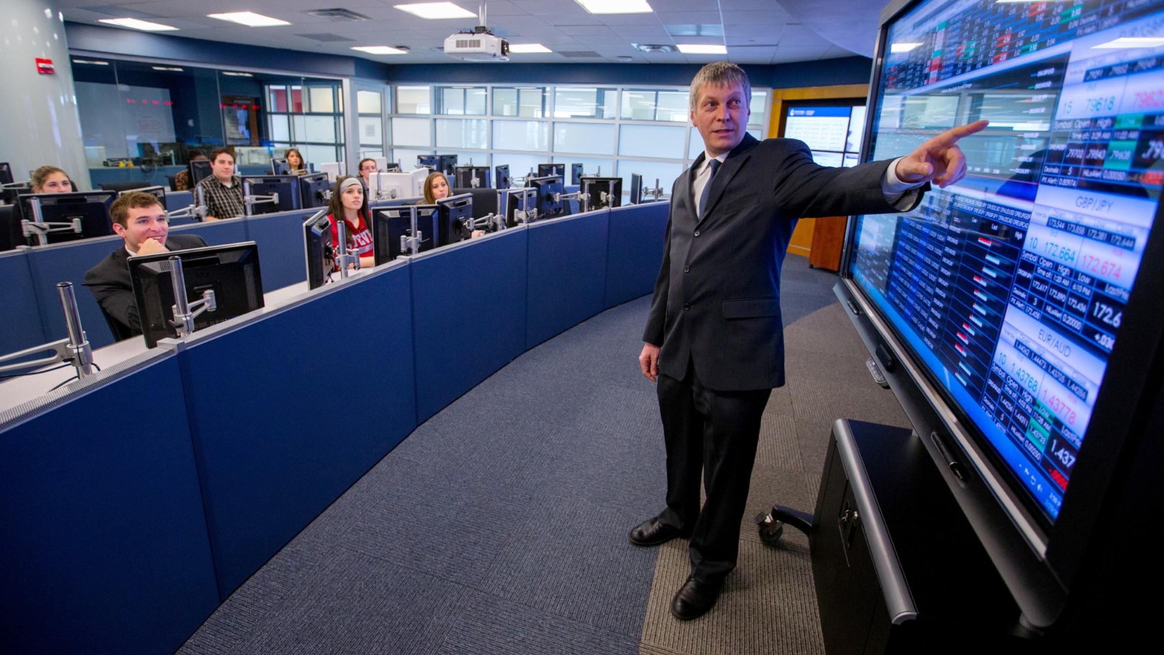 A male professor points to a screen readout of financial data as a class watches in a high-tech finance lab.